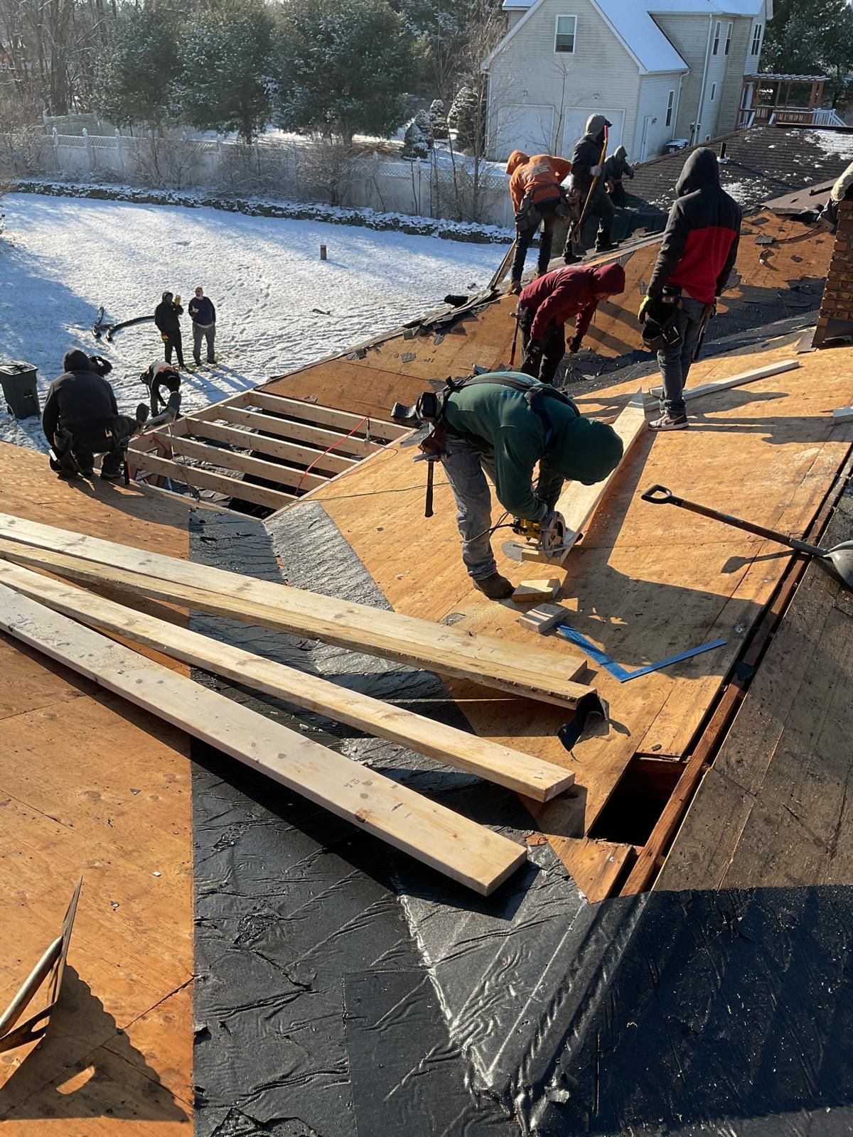 Workers in winter gear remove old roofing and install new plywood sheets on a snowy, sloped roof.