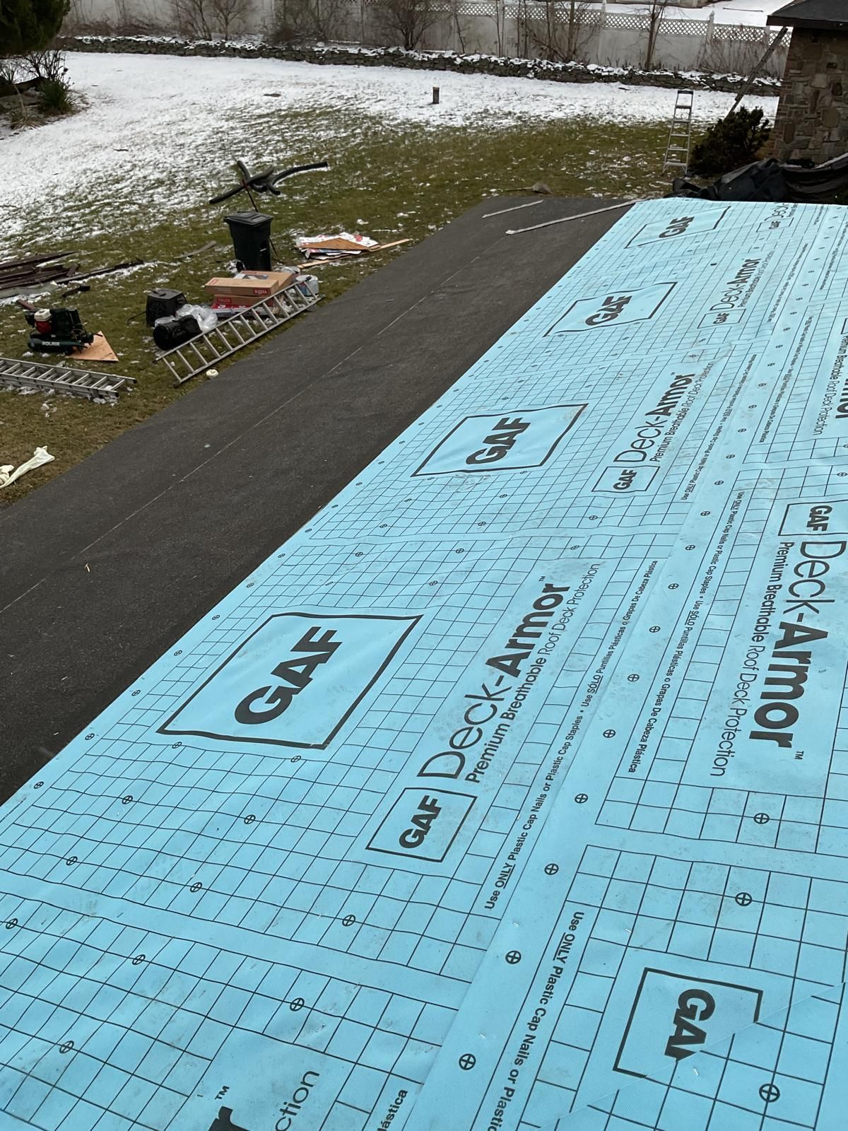 A roof under renovation, covered with blue GAF Deck-Armor underlayment, with a snowy backyard visible below.