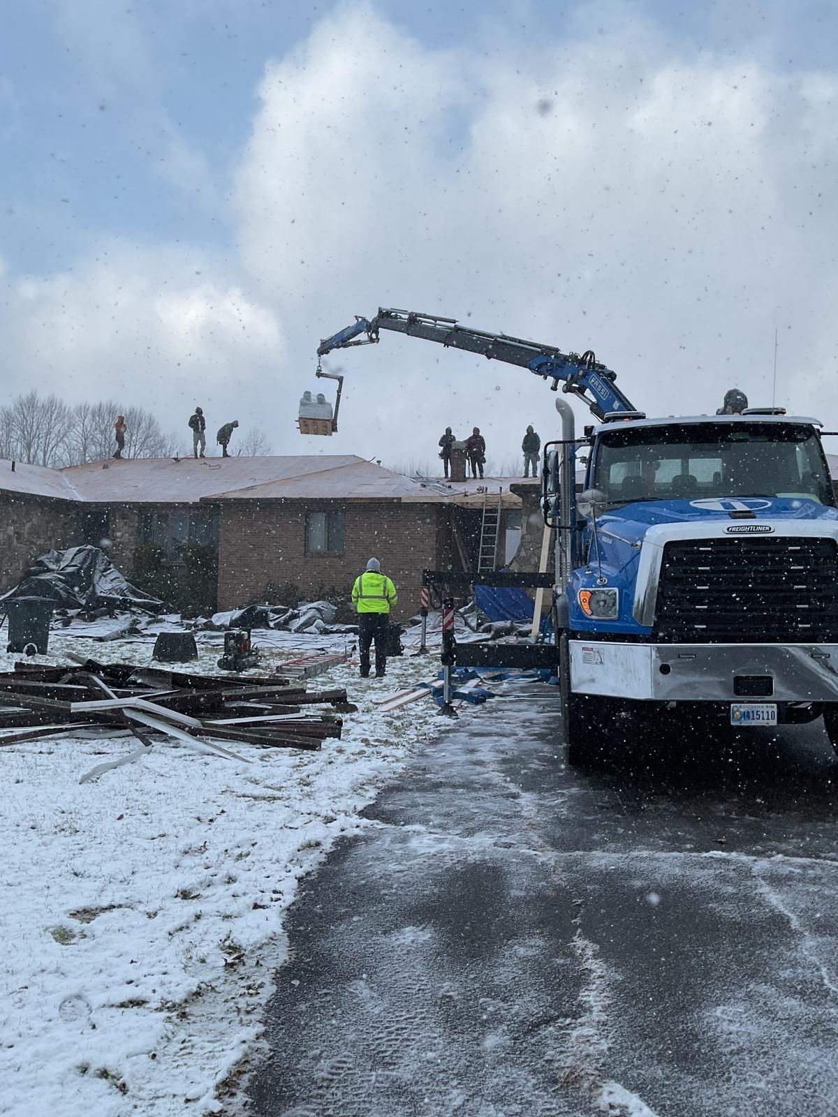 A blue utility truck with a crane arm lifting a basket to workers on a snowy residential roof.