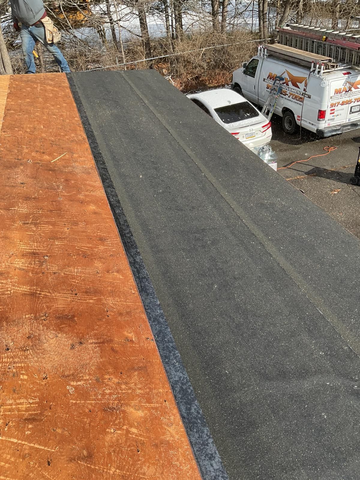 A partially installed roof showing bare plywood next to a strip of dark, granulated roofing material above a parking lot.