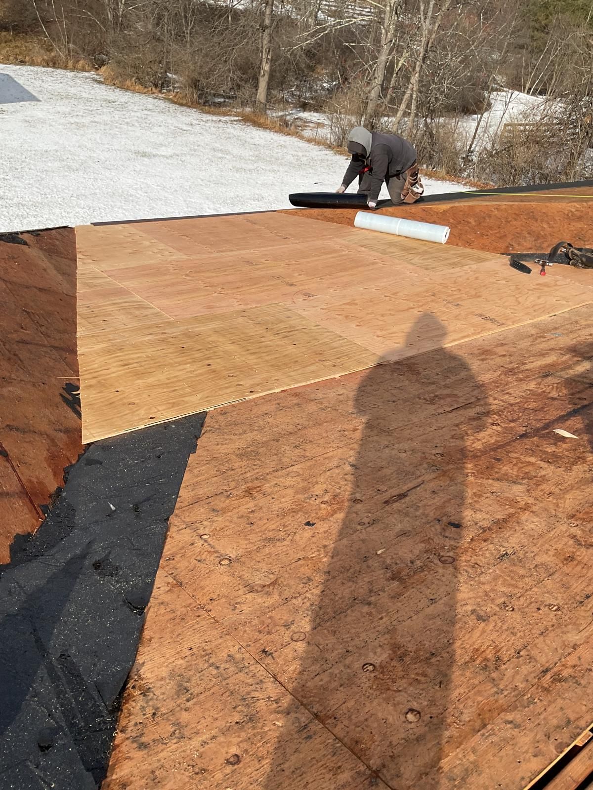 A person in a hooded jacket installs dark roofing underlayment onto wooden roof decking outdoors.
