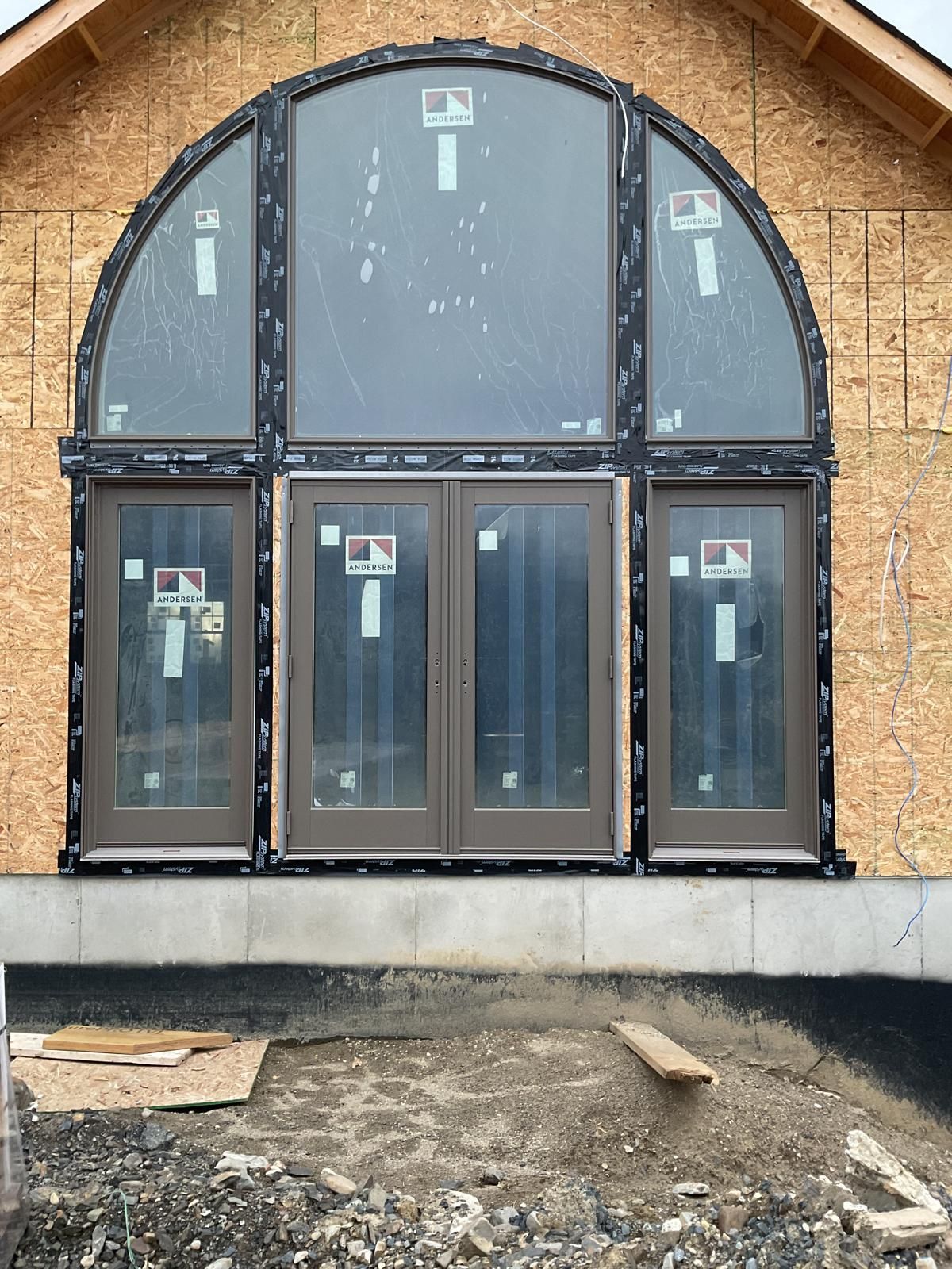 A large, brown-framed arched window assembly installed in the wooden exterior wall of an unfinished house construction.