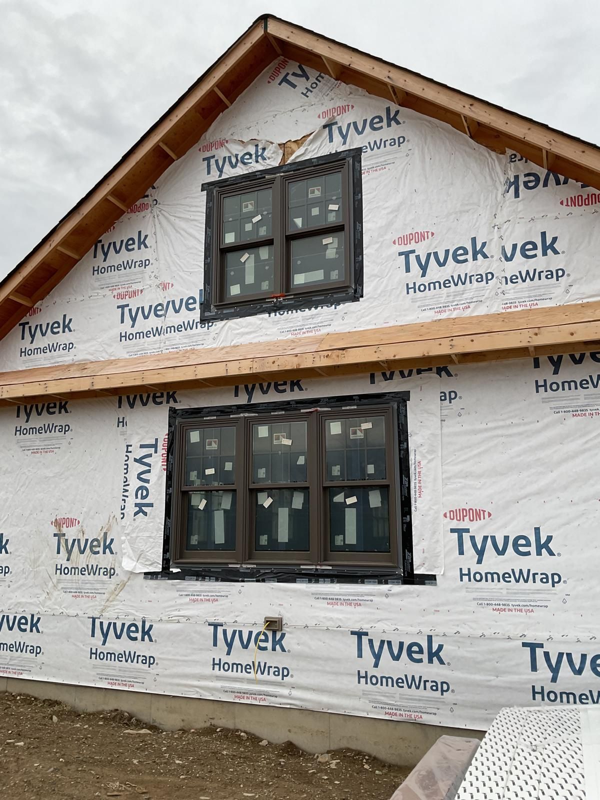 Exterior of a wood-framed house under construction featuring installed windows and white Tyvek house wrap.