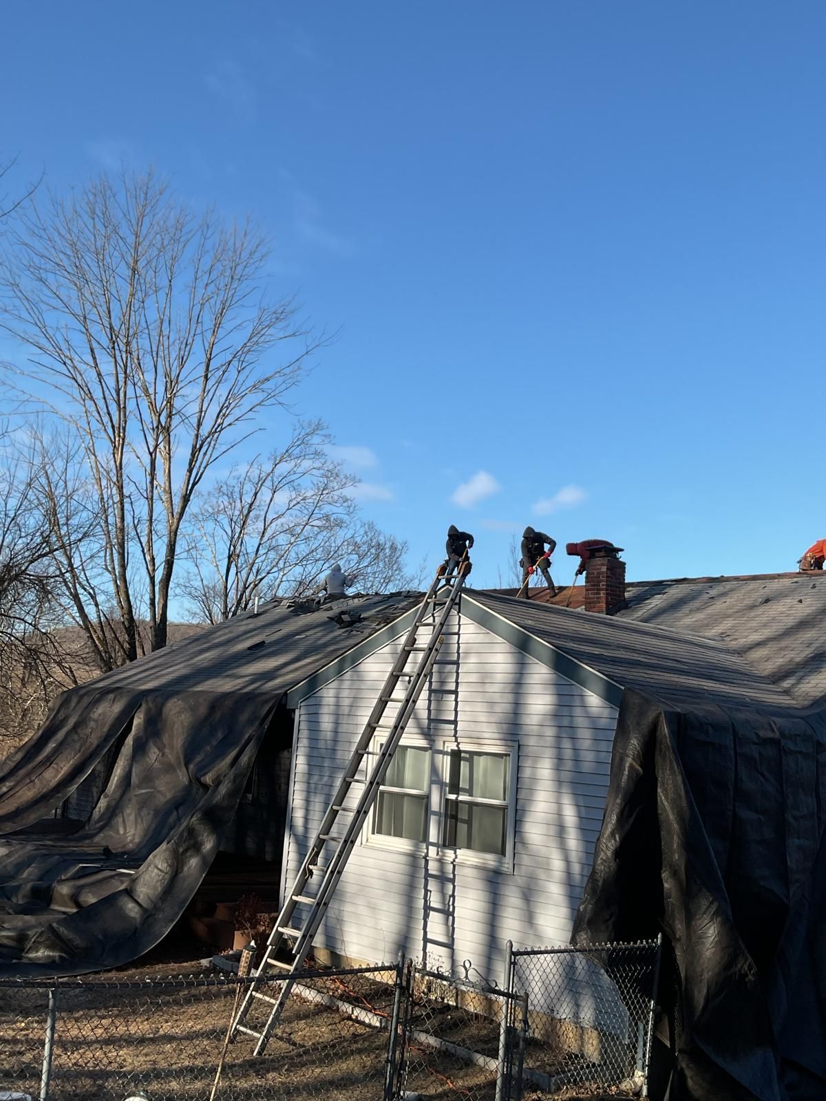 Two workers stand on the roof of a small house covered in black tarps, accessed by a long metal extension ladder.
