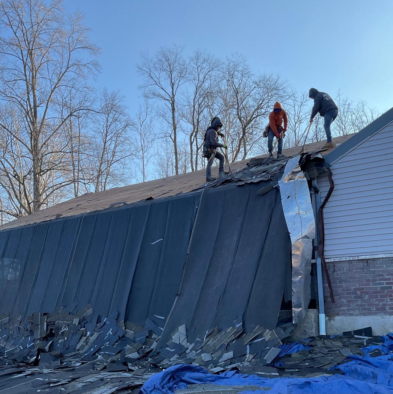 Three roofers remove old black shingles from a house roof on a sunny day.