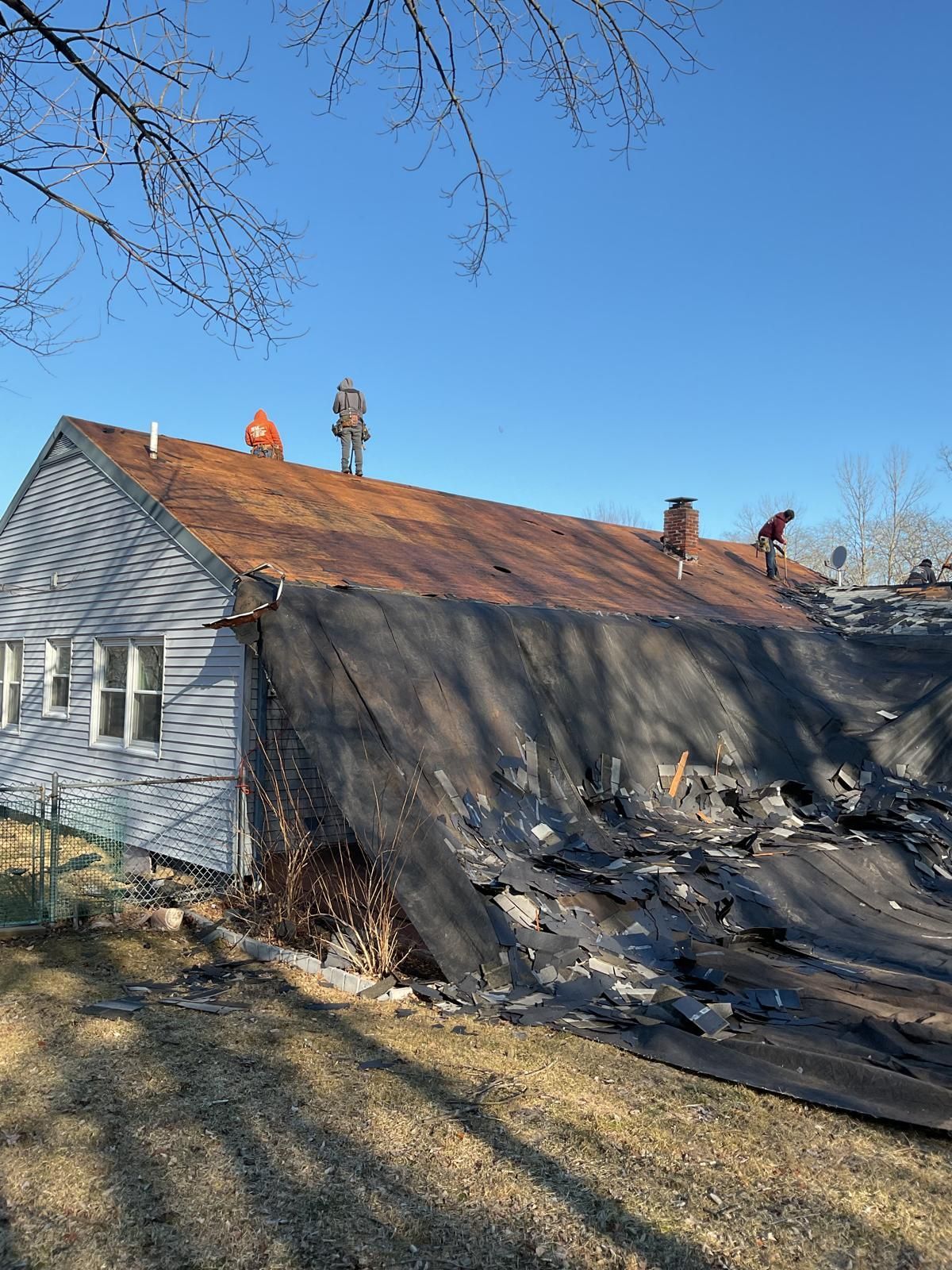 Construction workers replace the roof of a house, with old shingles piled on a tarp on the ground below.