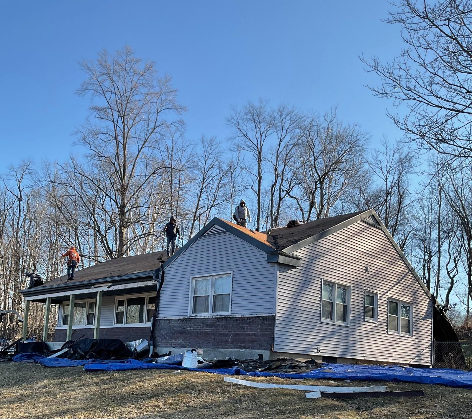 Workers on the roof of a light-colored, single-story house during a renovation, surrounded by bare trees under a blue sky.