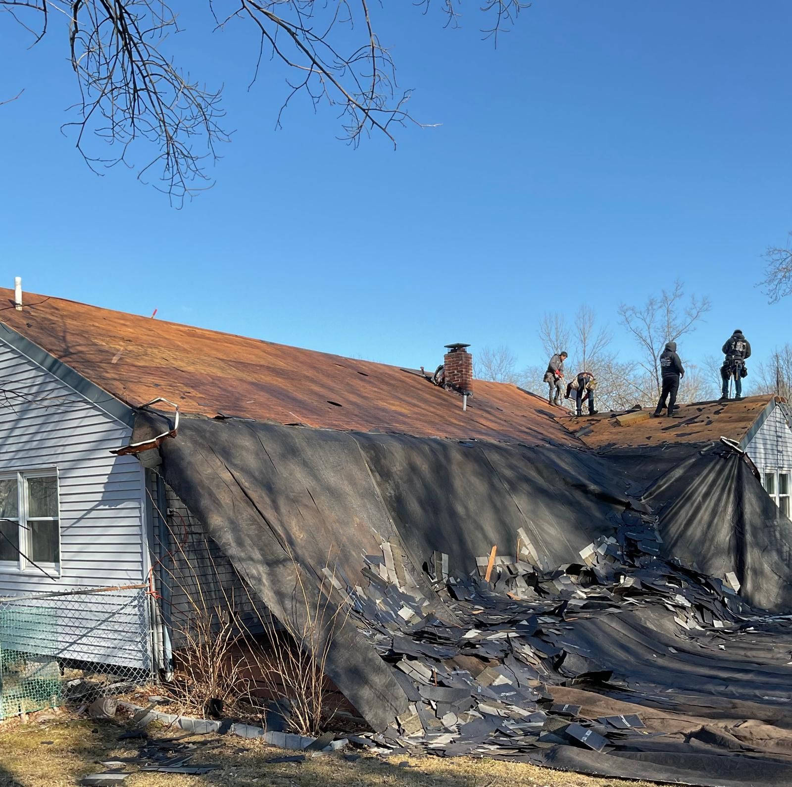 Four workers on a house roof stripping away damaged, fire-scorched shingles under a clear blue sky.