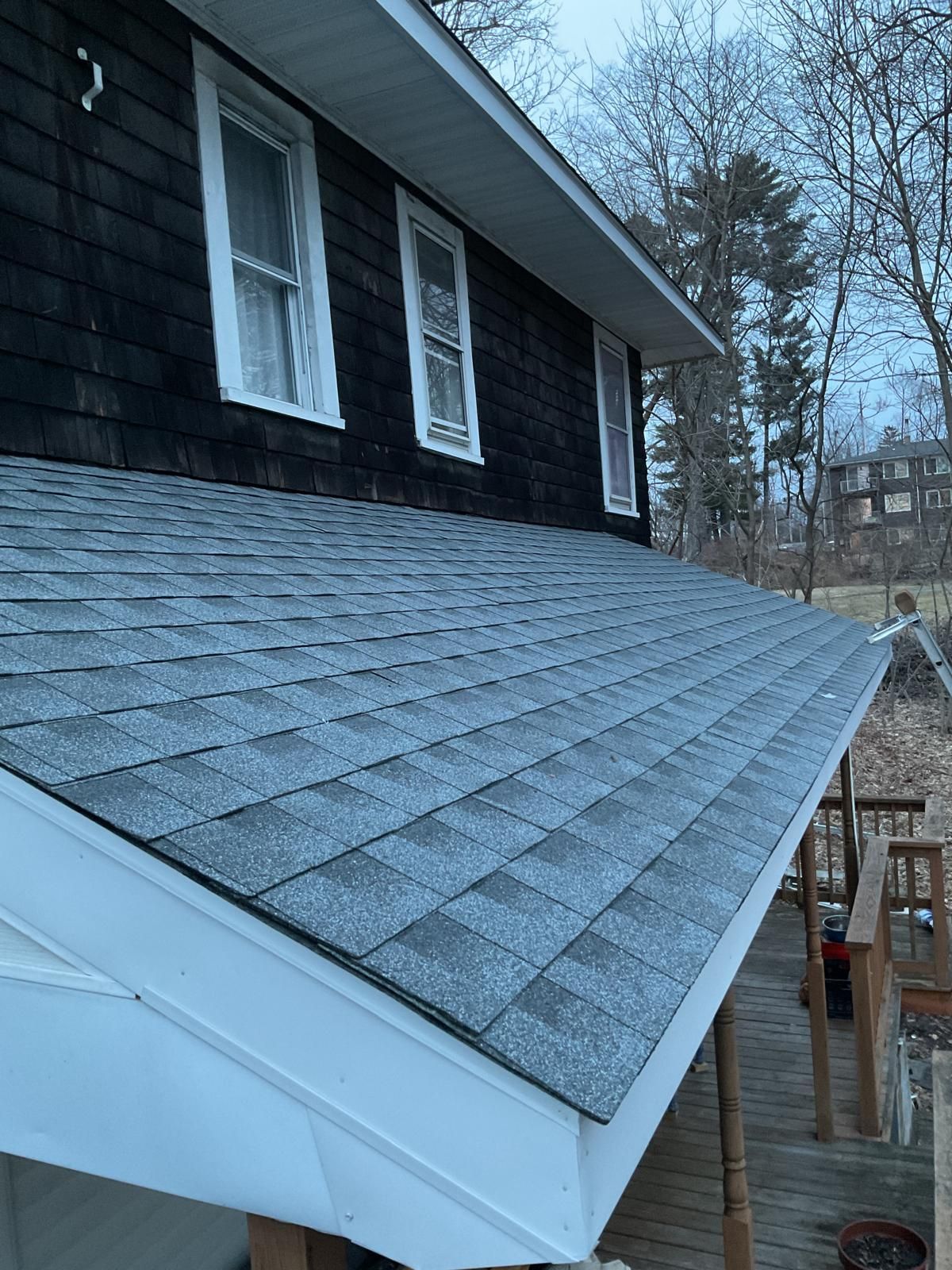 A close-up of a house roof featuring gray shingles, white trim, and dark siding with two windows, viewed from a deck.