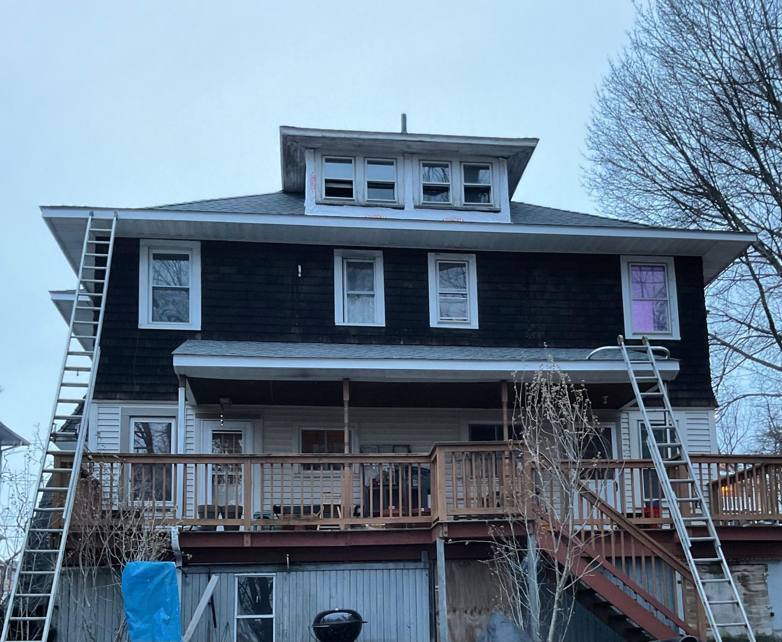 A two-story dark-sided house with a dormer window, wooden deck, and two ladders leaning against the exterior walls.