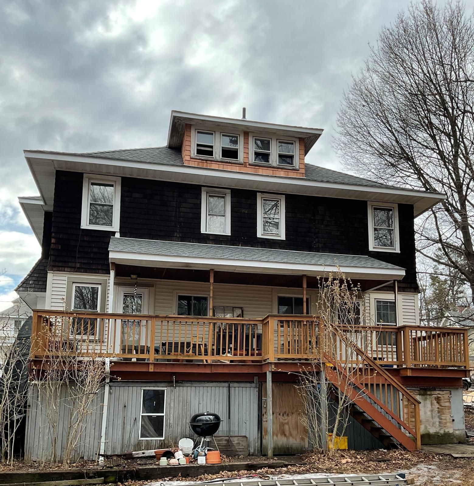 A two-story house with black siding, a white lower level, a large wooden deck, a staircase, and a roof dormer.