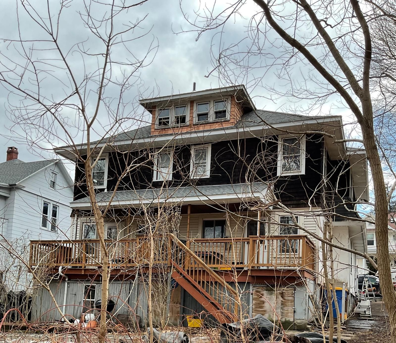 A weathered, two-story house with dark siding, a wooden deck, and a small attic dormer stands behind bare trees.