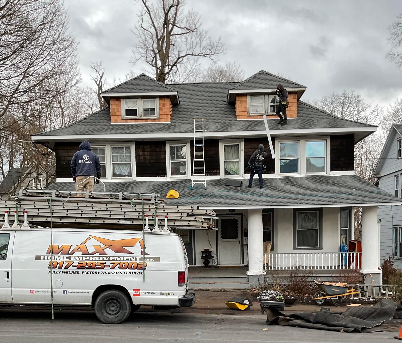 Workers on a roof, preparing to replace shingles, with a van labeled Max Asphalt parked in front of a two-story house.