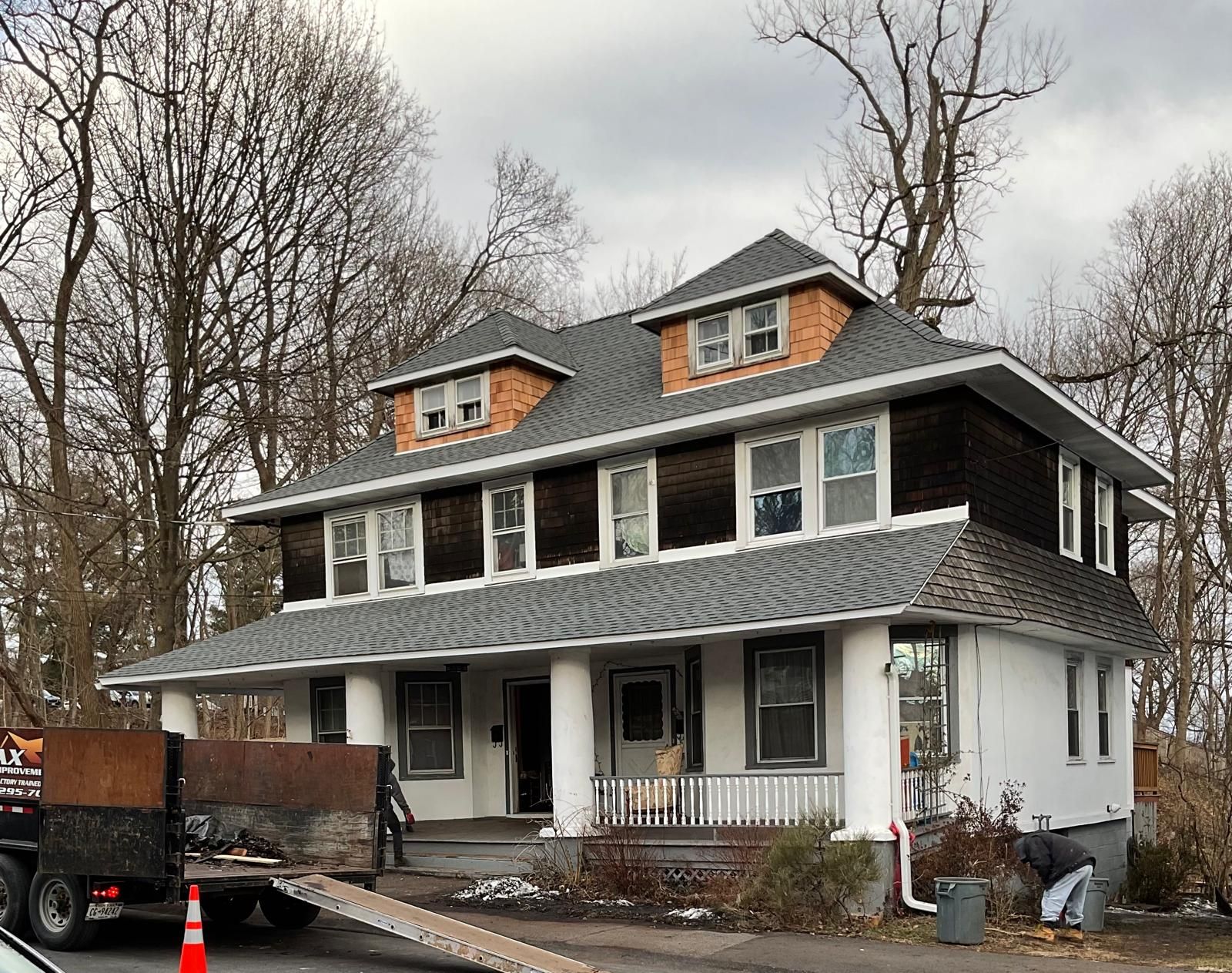 A two-story house with dark shingle siding, a white first floor, and a front porch, viewed from a street with a truck.