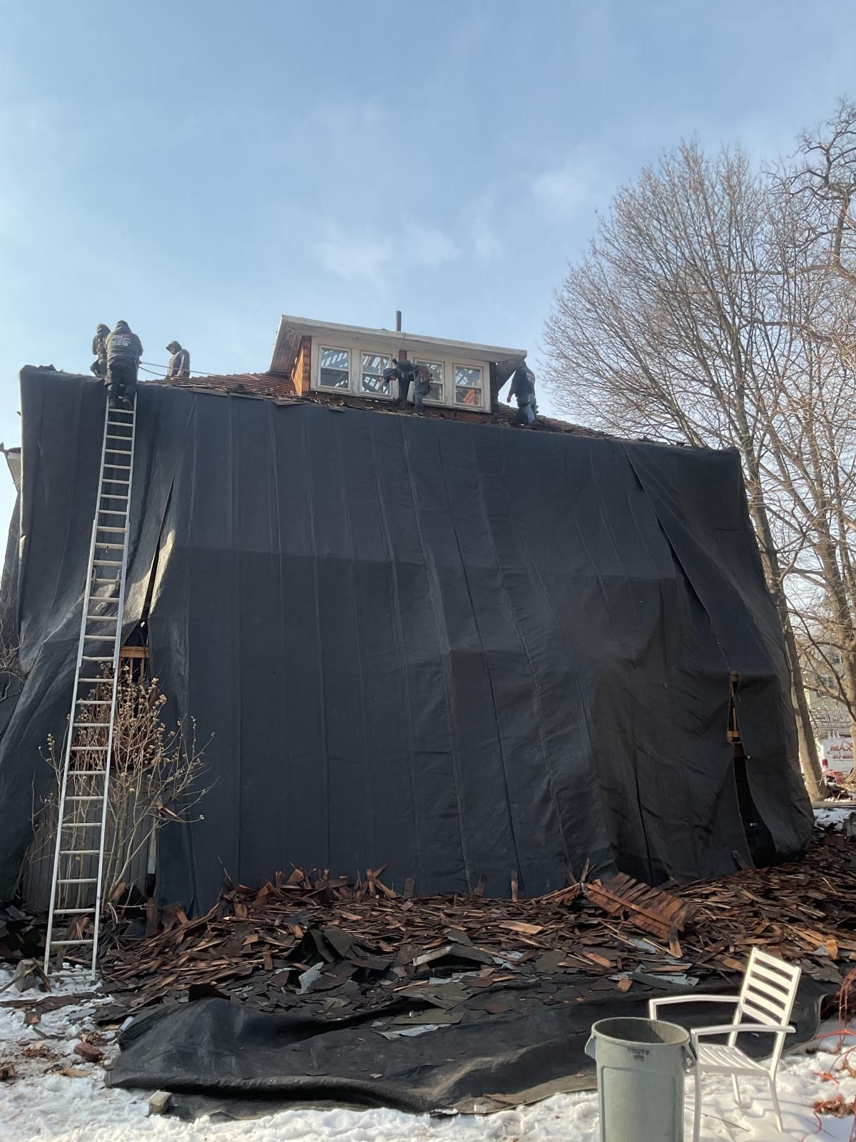 Workers on the roof of a house undergoing repairs, with a large black tarp covering the wall and debris on the ground.