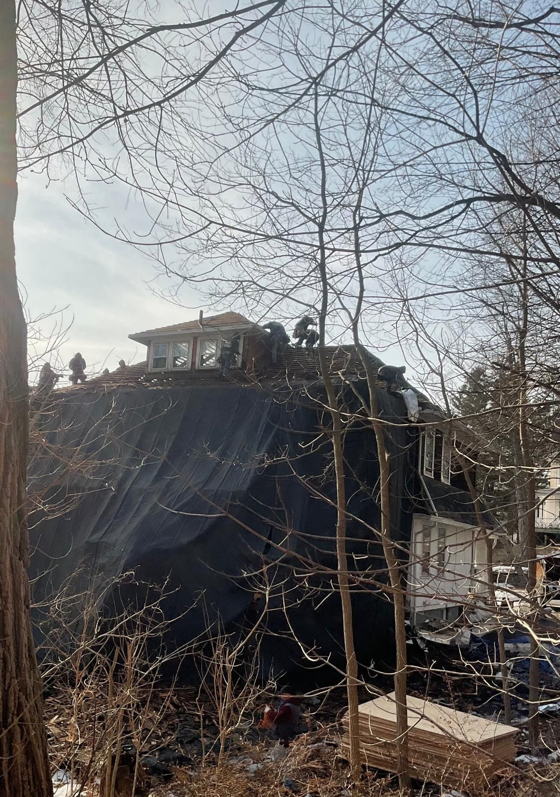 Workers on the roof of a house partially covered by a large black tarp, viewed through surrounding bare trees.