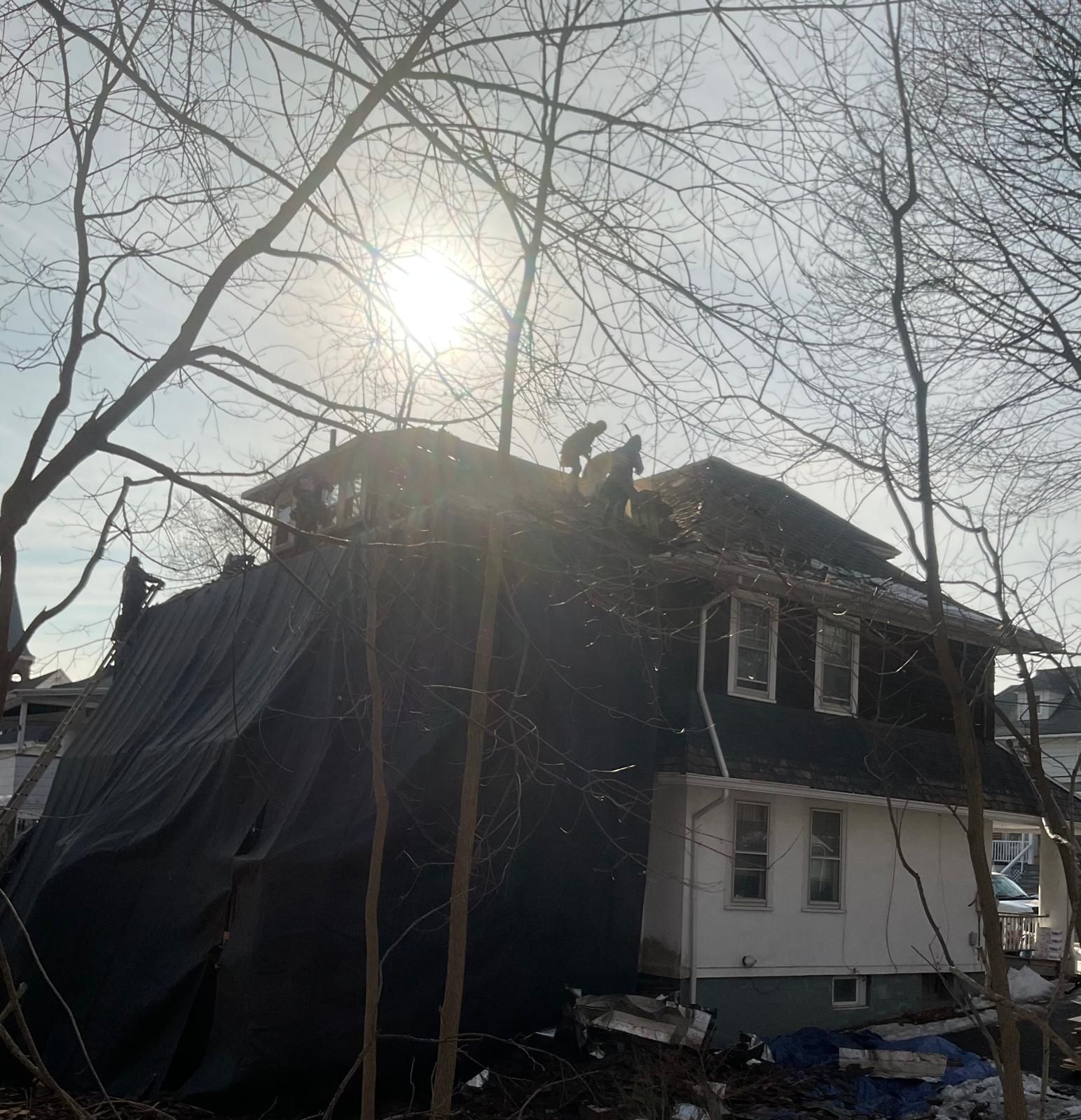 Workers on the roof of a two-story home, partially covered by black tarps, during a sunny day with bare trees in front.