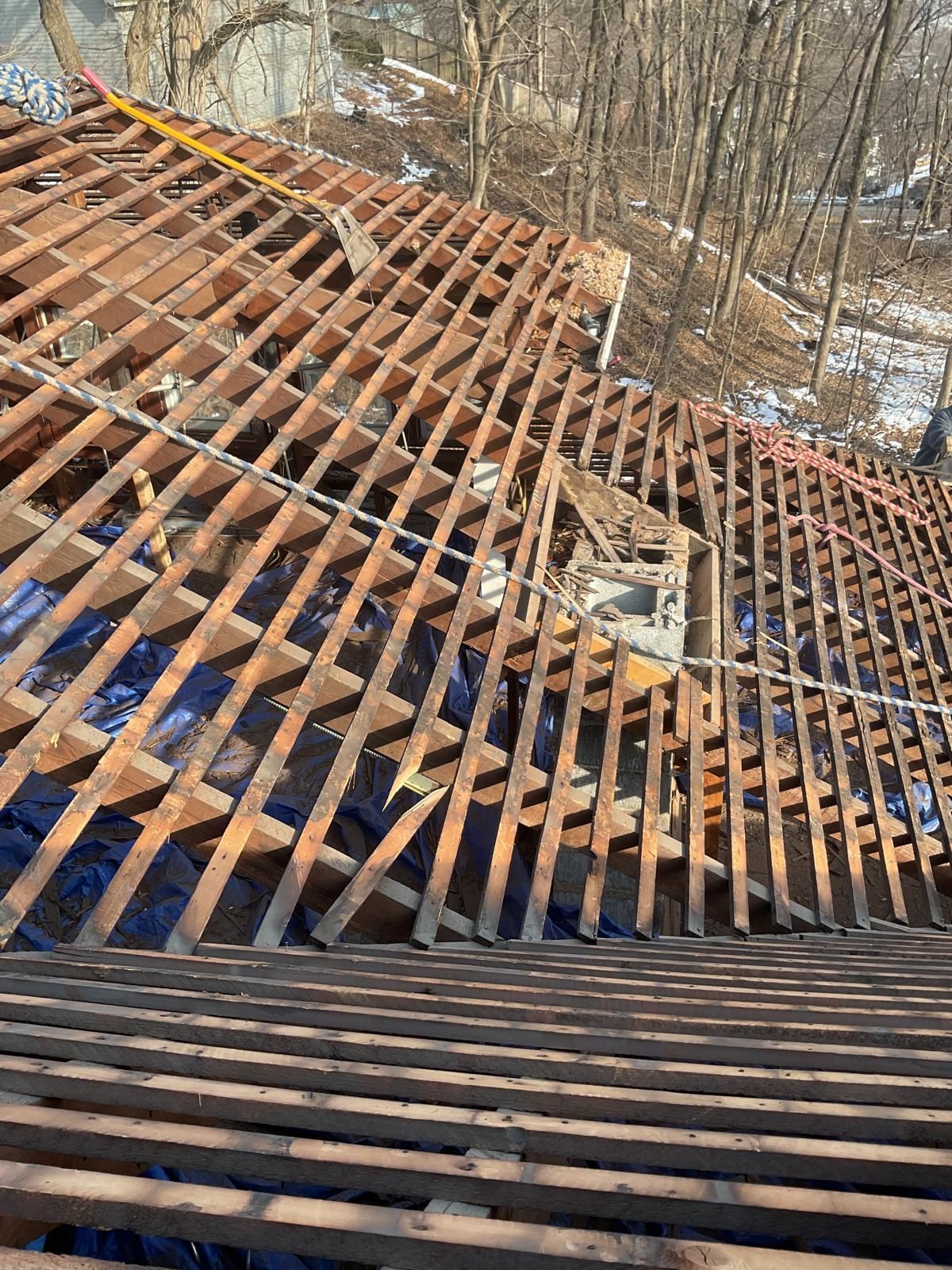 An overhead view of a roof structure under construction, showing wooden rafters and horizontal laths over a wooded area.