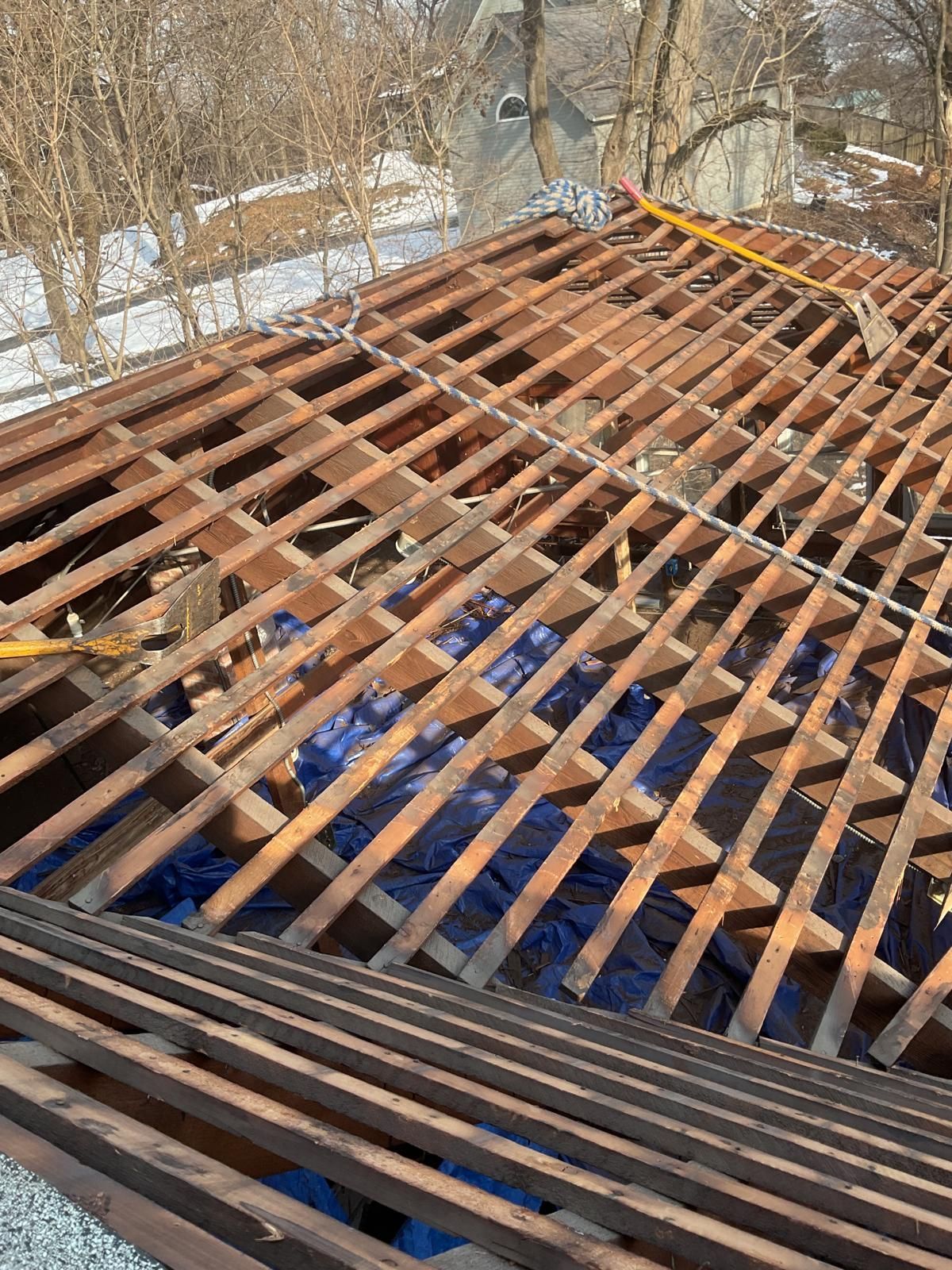 A view of a roof under construction, showing bare wooden rafters and slats with a blue tarp visible underneath.