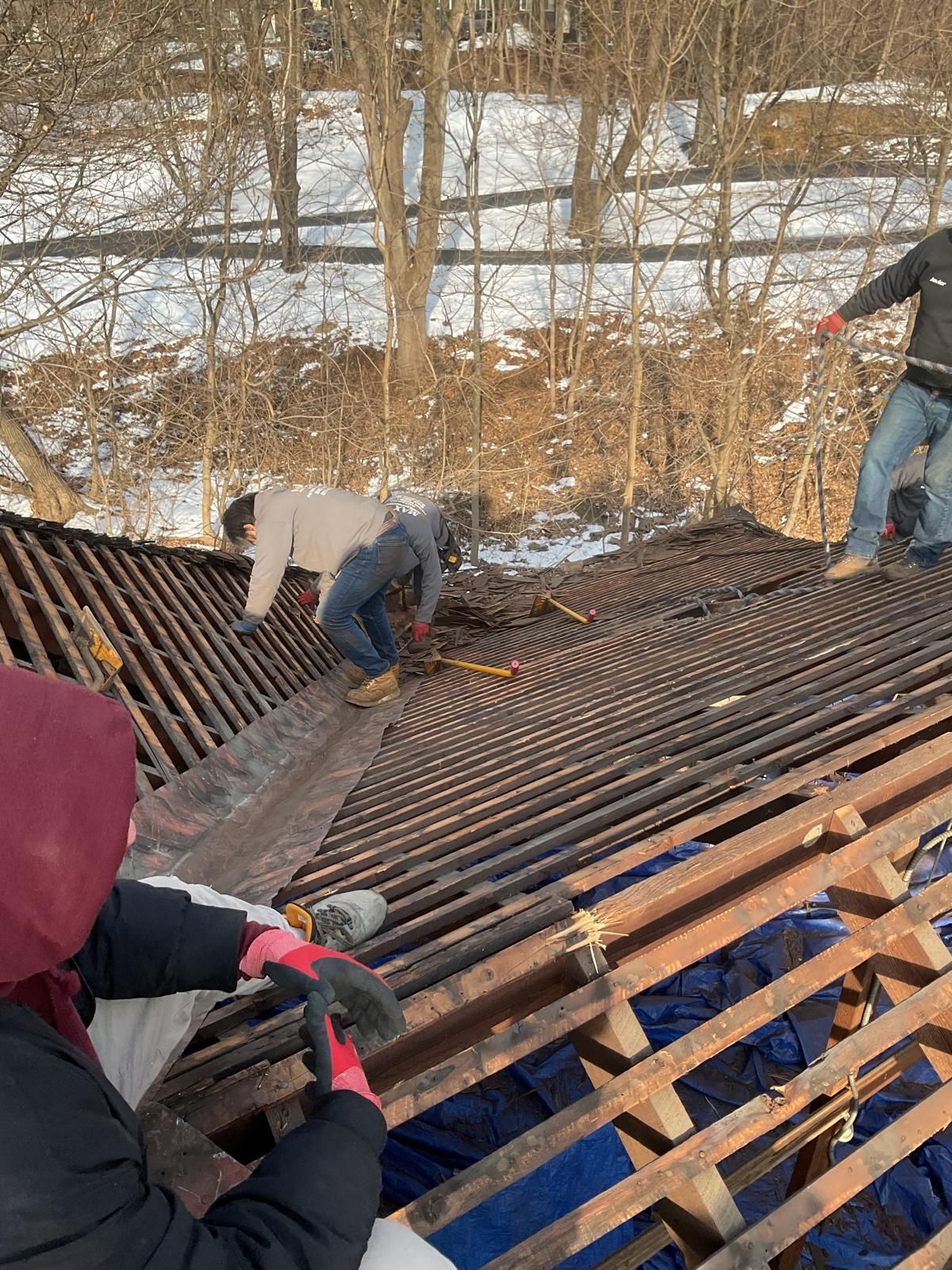 Three workers in winter clothing remove old roofing materials from the wooden frame of a roof, with trees and snow nearby.