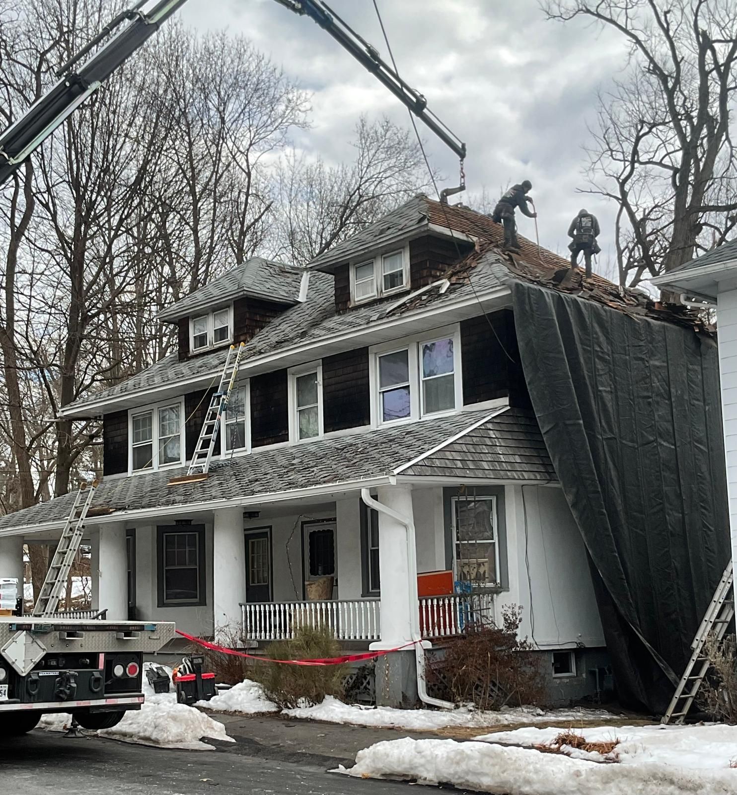 Workers on the roof of a two-story house use a crane to remove shingles, with a large tarp draped along the side.