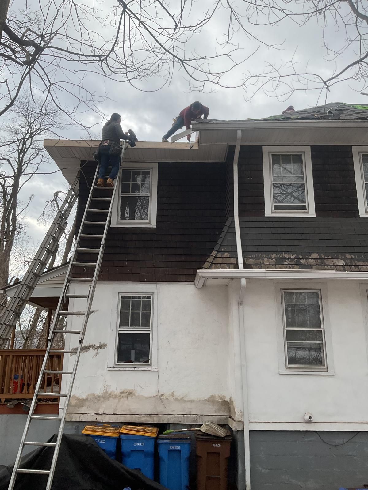 Two people work on the roof and gutters of a two-story home with white and dark brown exterior walls.