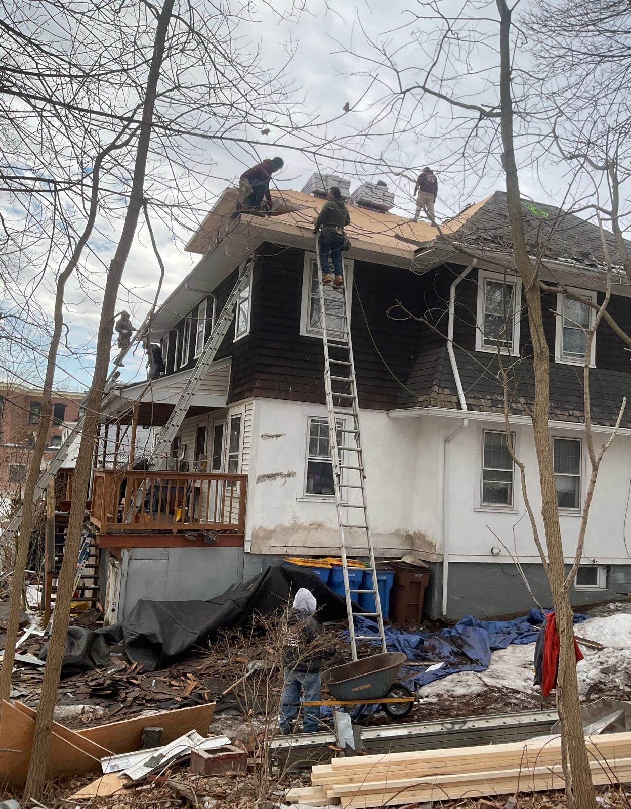 Workers replace a roof on a two-story home, with some on the roof and one climbing a ladder near a ground-level worker.