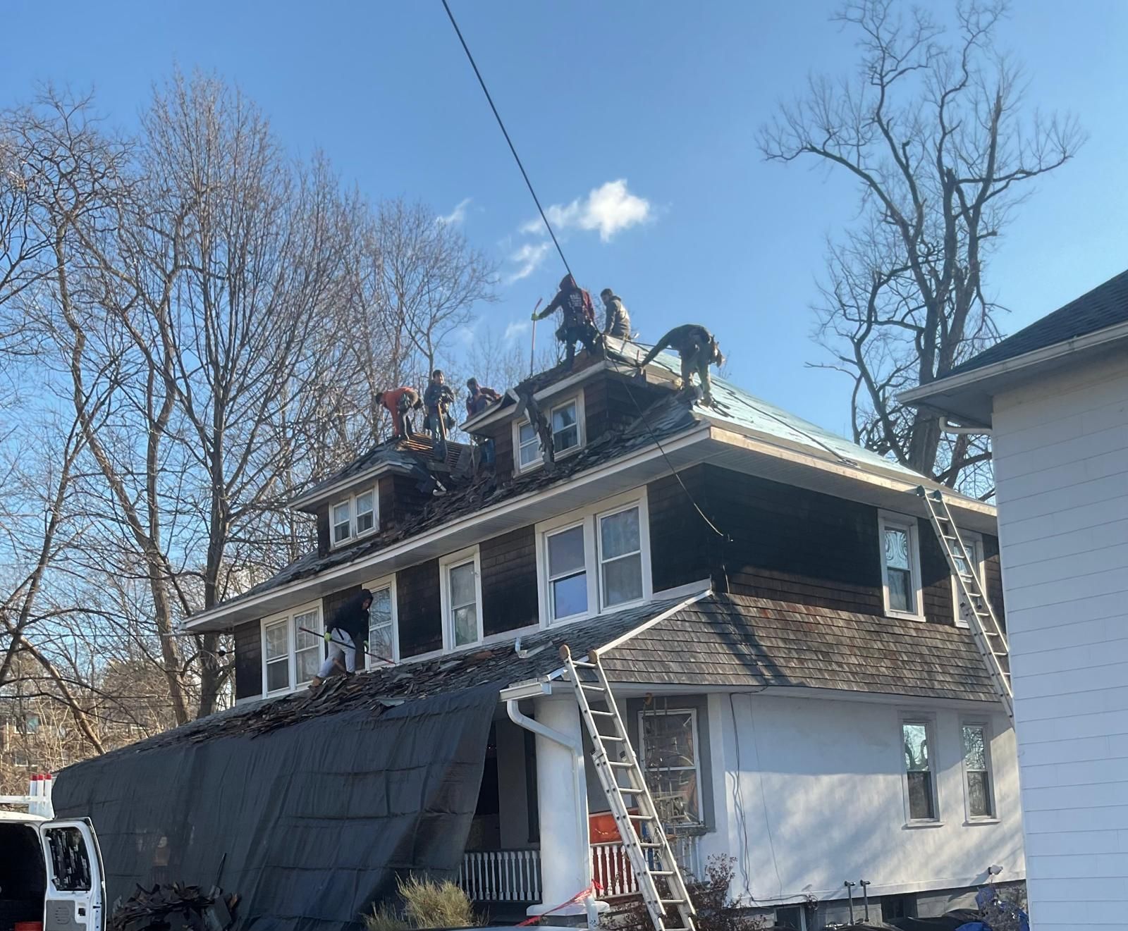 A team of roofers works on the top of a two-story house with a white exterior, using ladders and protective tarping.