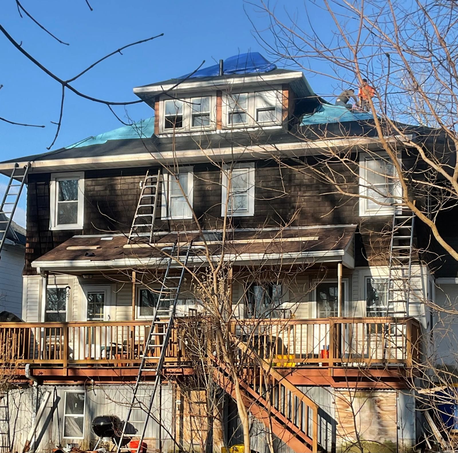 Workers repair the roof of a large, multi-story house covered in blue tarps, featuring a deck and multiple ladders.