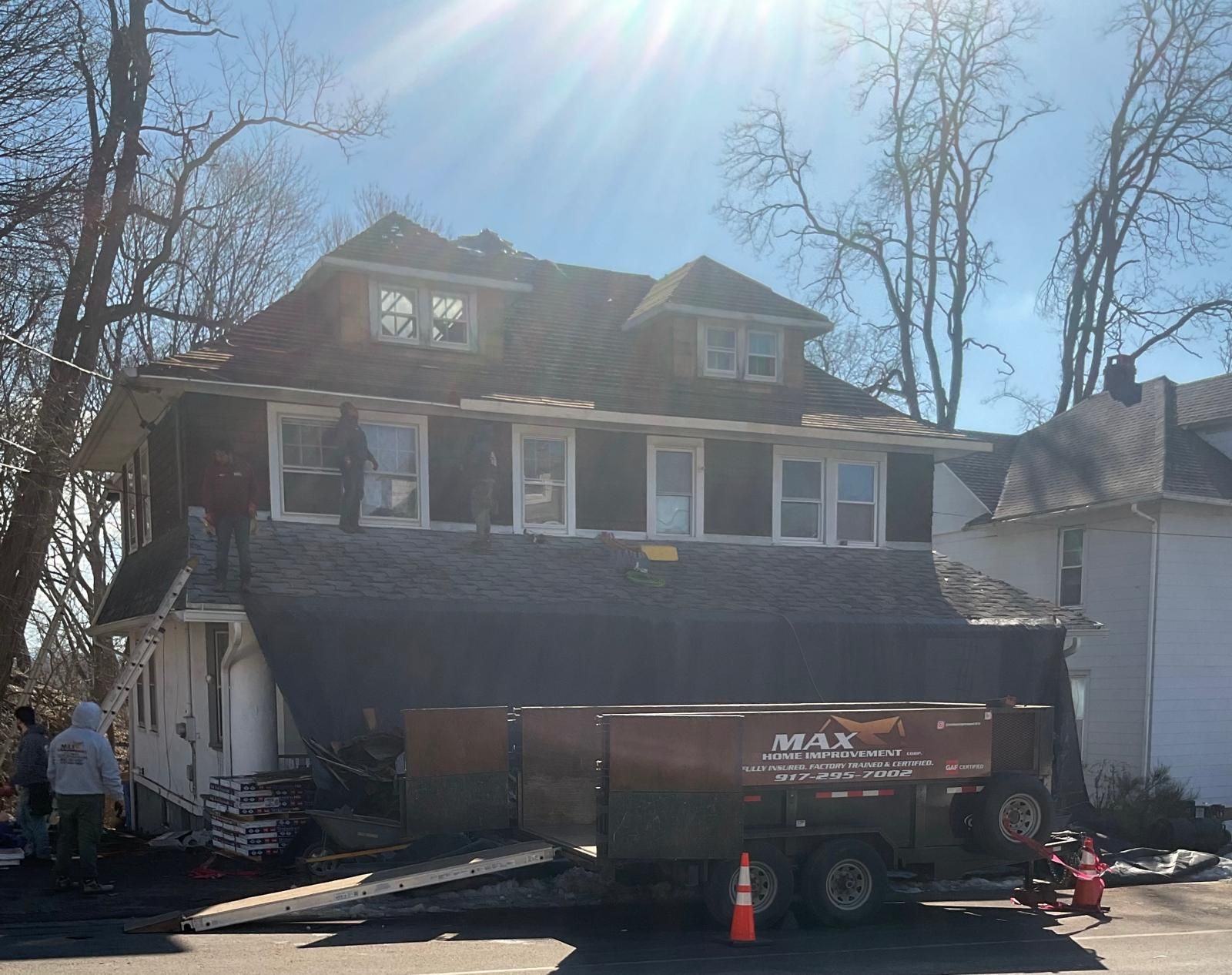 Workers removing old roofing shingles from a two-story home, with a dumpster trailer parked in the driveway.