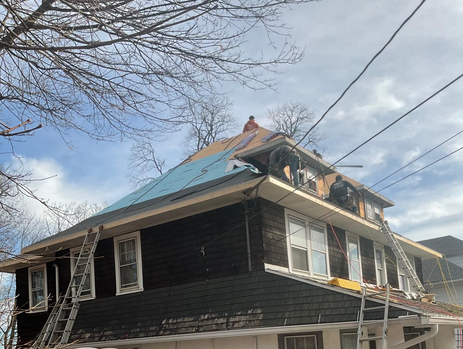 Workers are installing new roofing material on the upper level of a two-story house with dark siding.