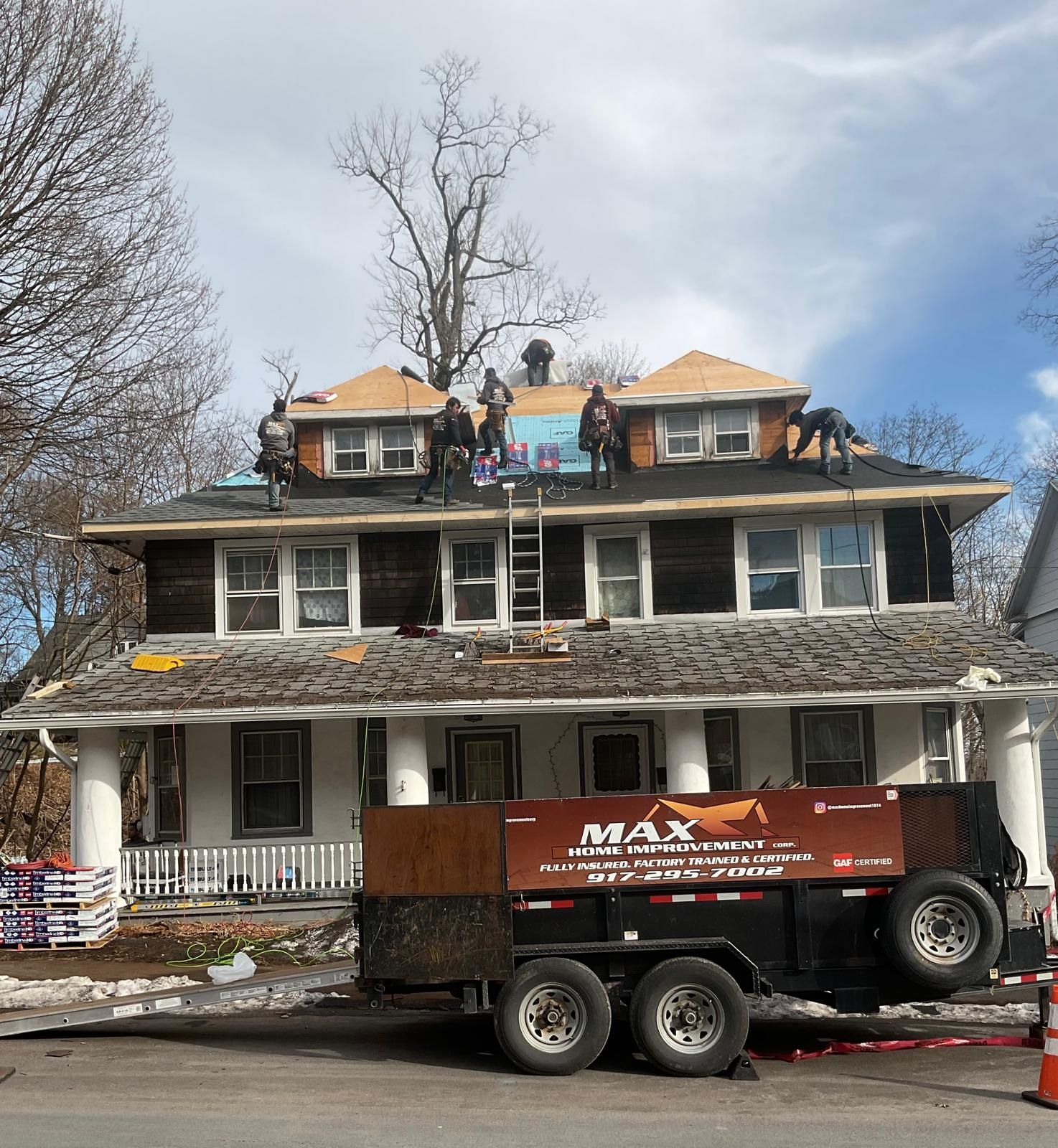 Several roofers work on the roof of a large, two-story house while a MAX dumpster trailer sits in the front yard.