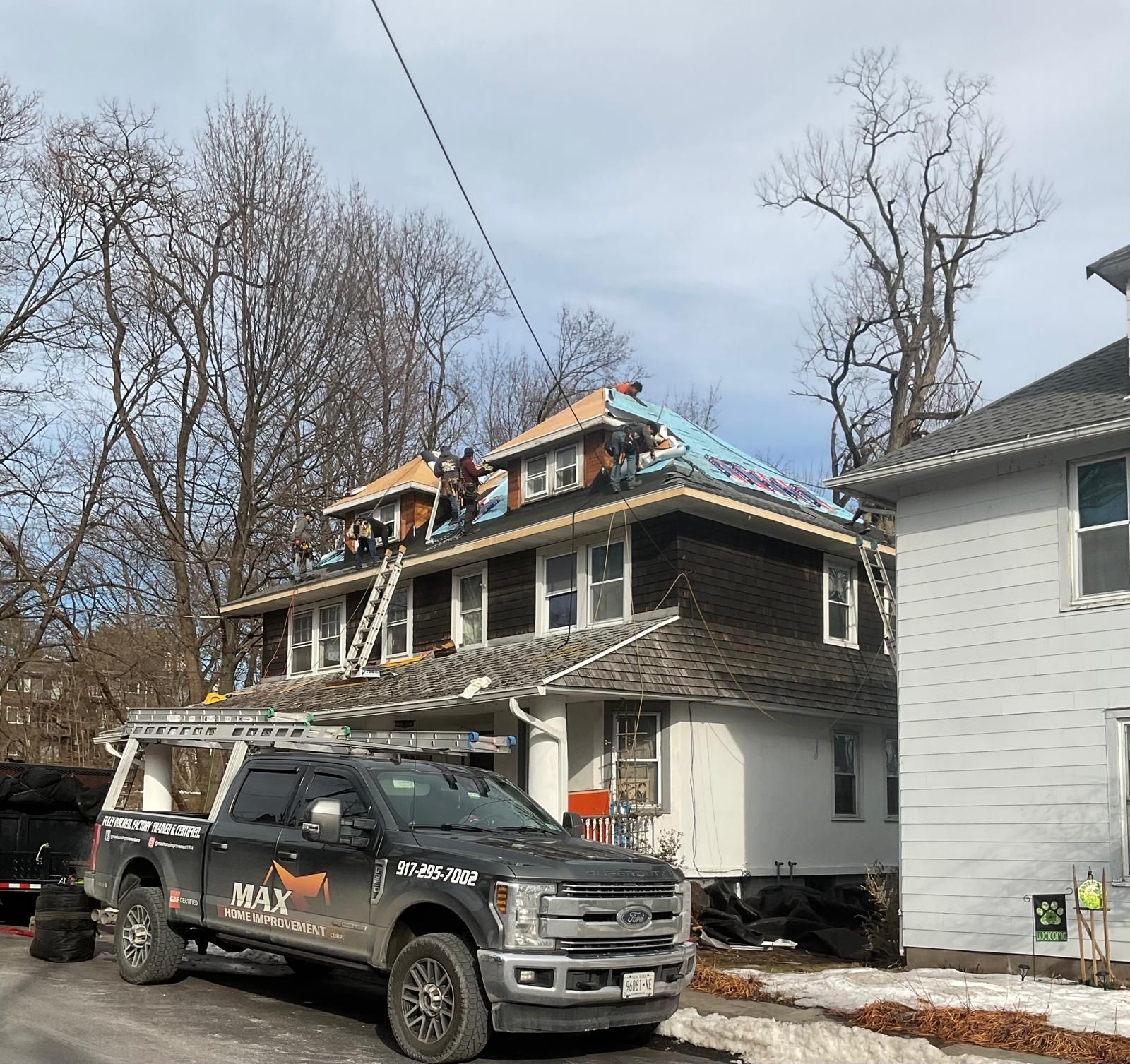 Roofers work on the shingled roof of a two-story home, with a Max Construction truck parked in the foreground.