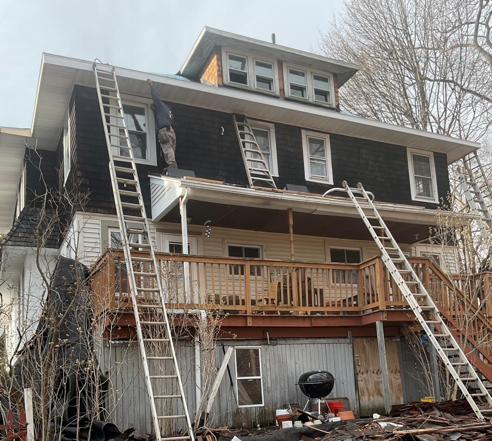 A three-story dark house undergoing exterior renovations with two long ladders resting against the roof and balcony.