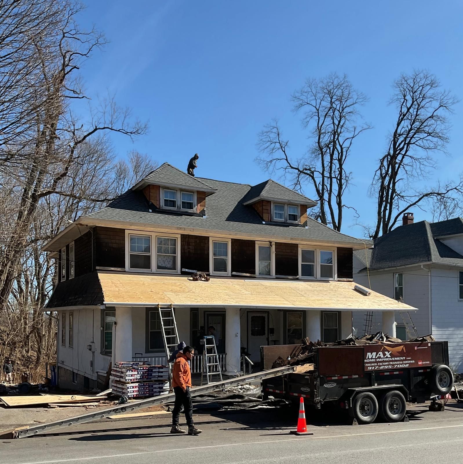 Workers repair the roof of a two-story home with a dumpster trailer parked in front on a sunny day.