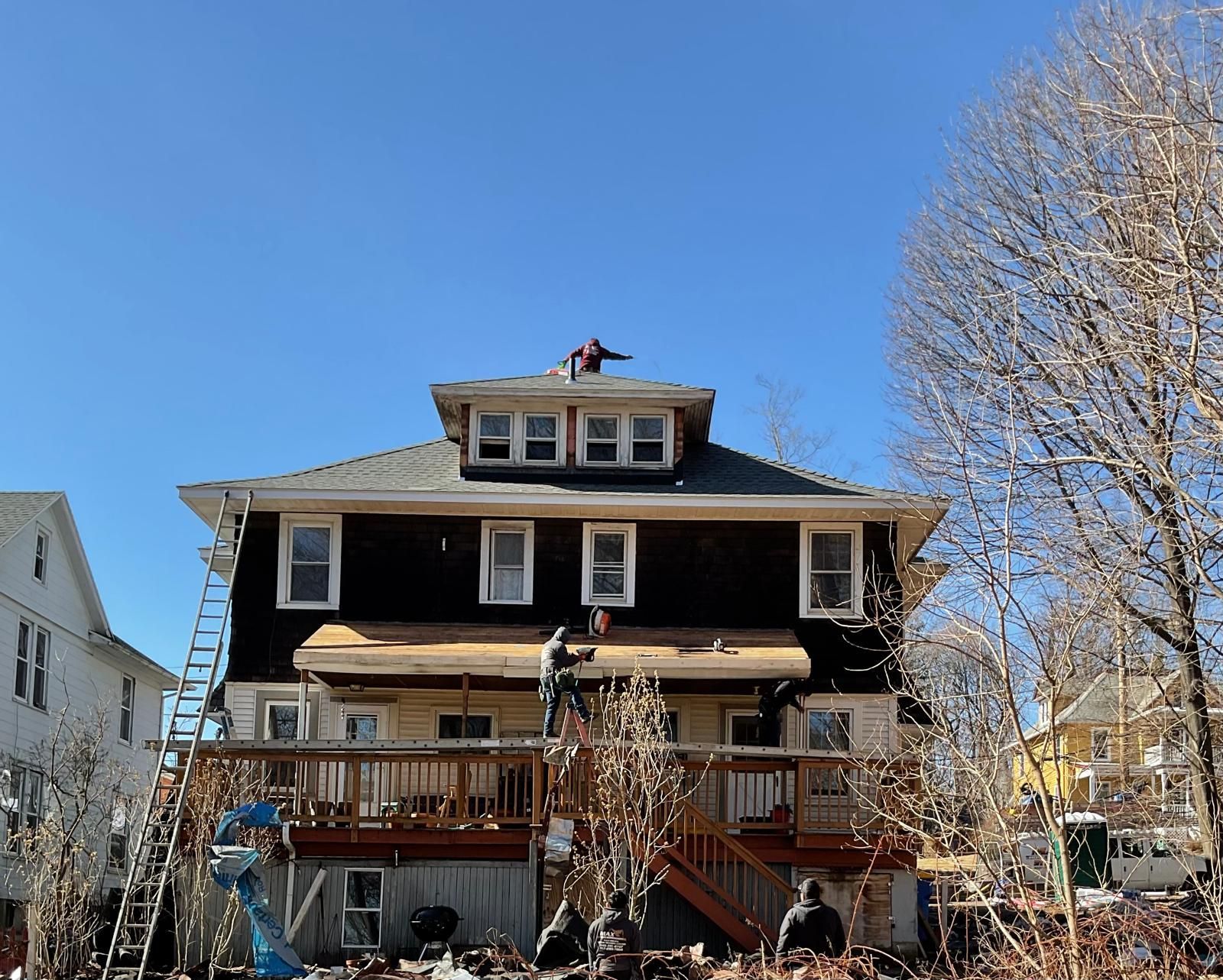 Workers renovate a multi-story house with a wooden deck and a black-shingled exterior under a clear blue sky.