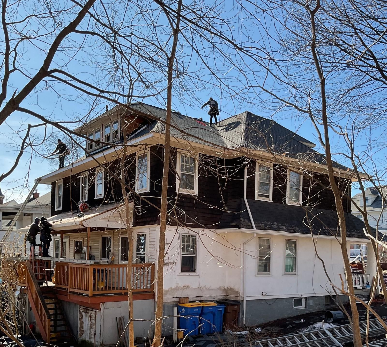 Contractors work on the roof and upper deck of a multi-story house with brown shingle siding and white lower walls.