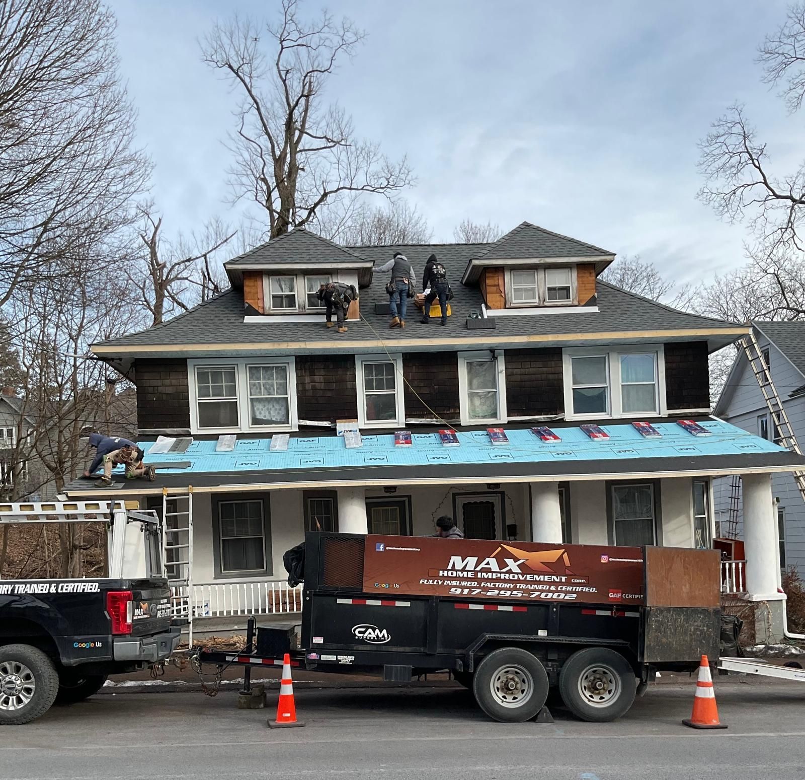 Construction workers repair the shingled roof of a house, with a parked trailer marked 