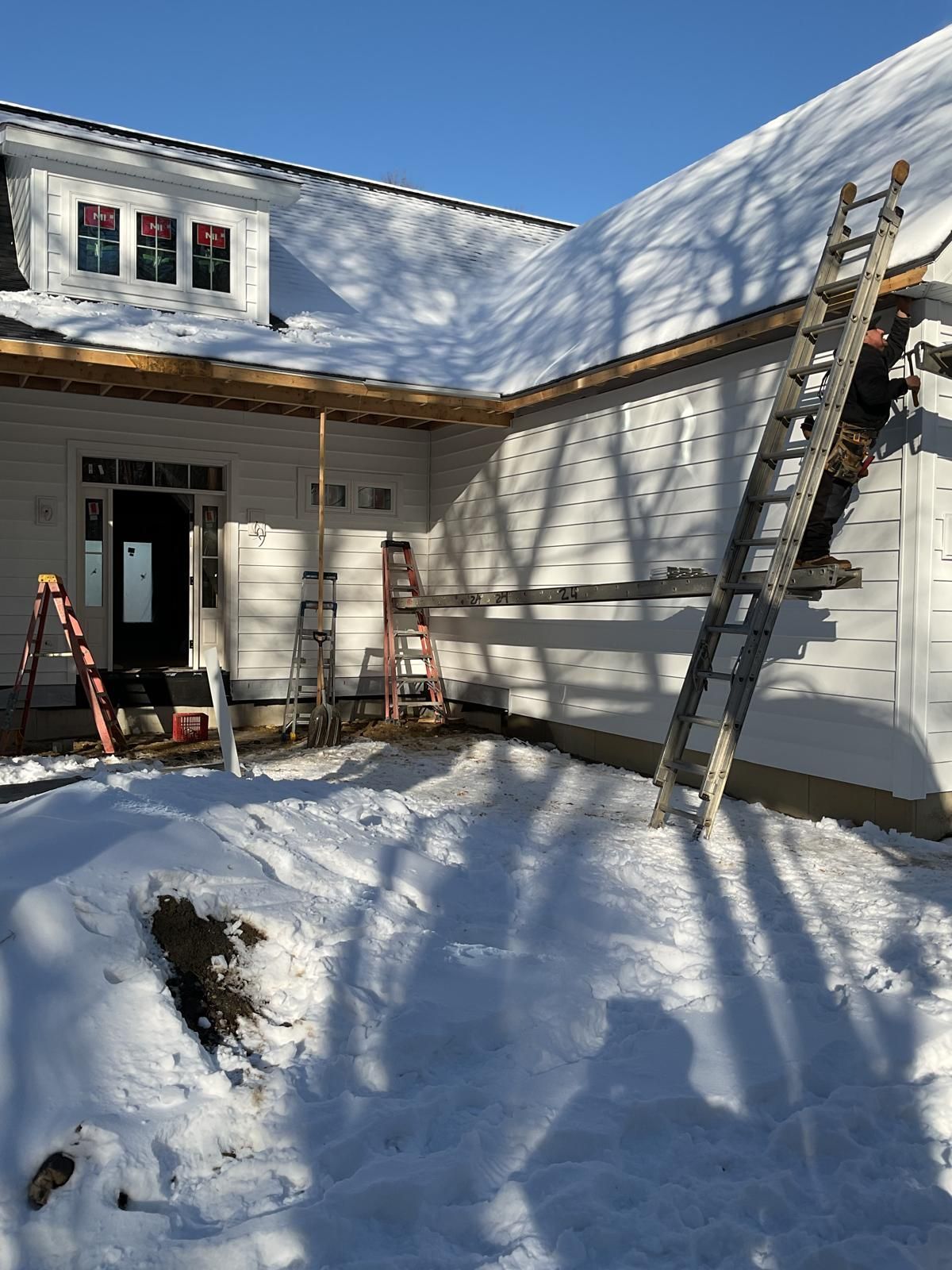 A worker on a ladder installs siding on a snowy, residential construction site.