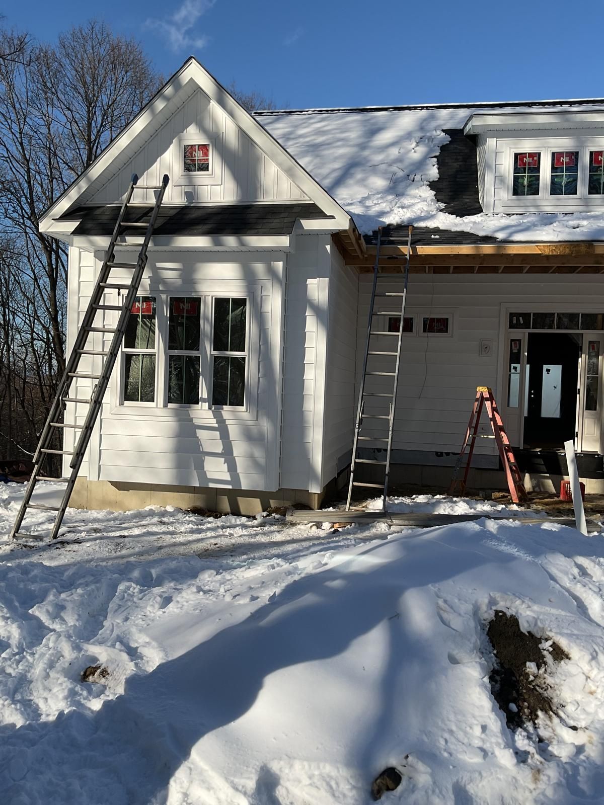 A house under construction with white siding and ladders against its exterior, set in a snowy landscape under a blue sky.