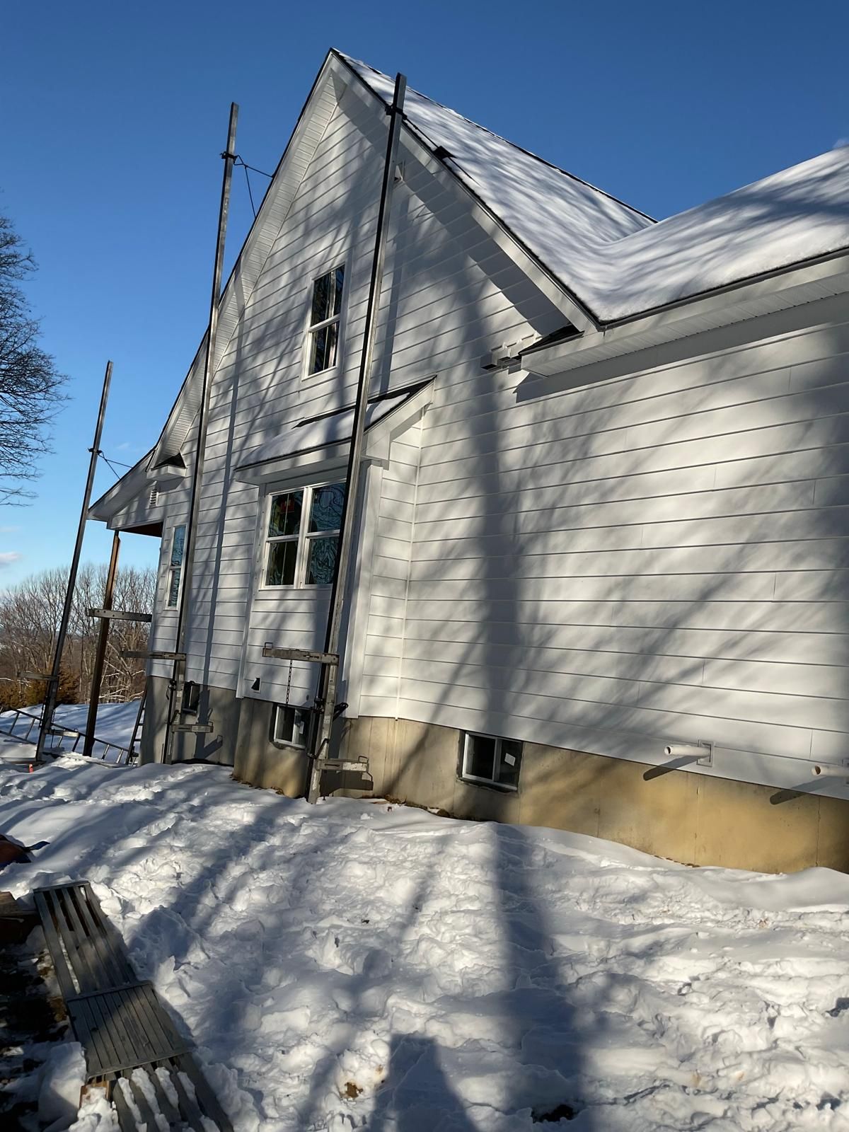 A two-story white house exterior under construction, with scaffolding attached, surrounded by snow on a sunny day.