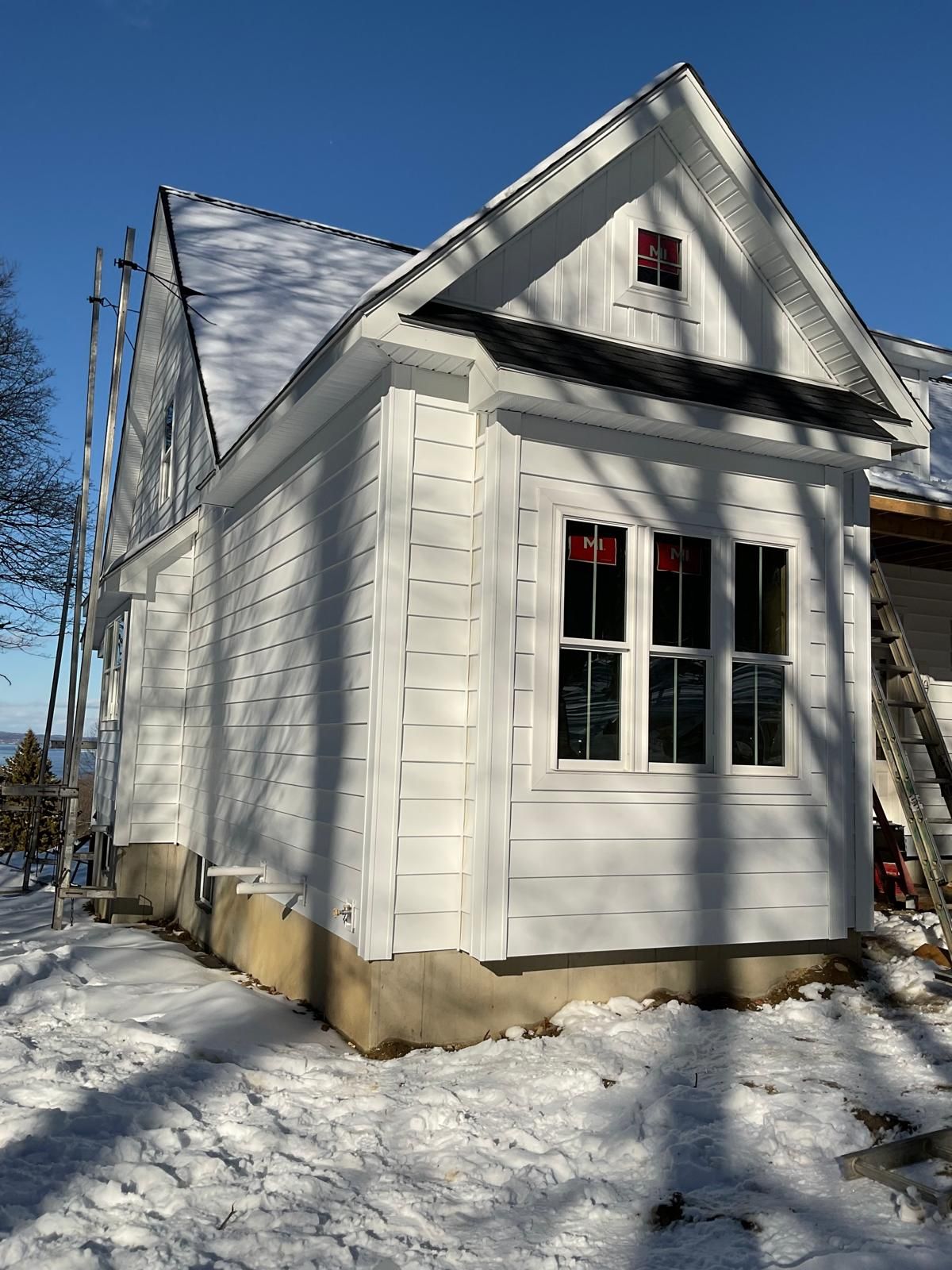 A partially constructed white-sided house addition with a large window and gable roof stands on snow-covered ground.