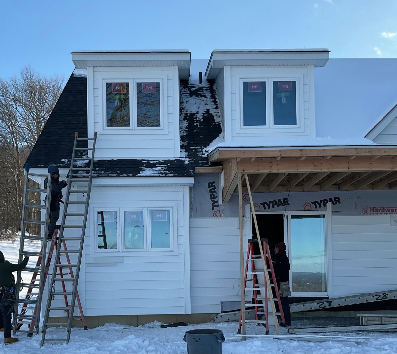 Workers use ladders to install siding on a white house under construction in a snowy landscape.