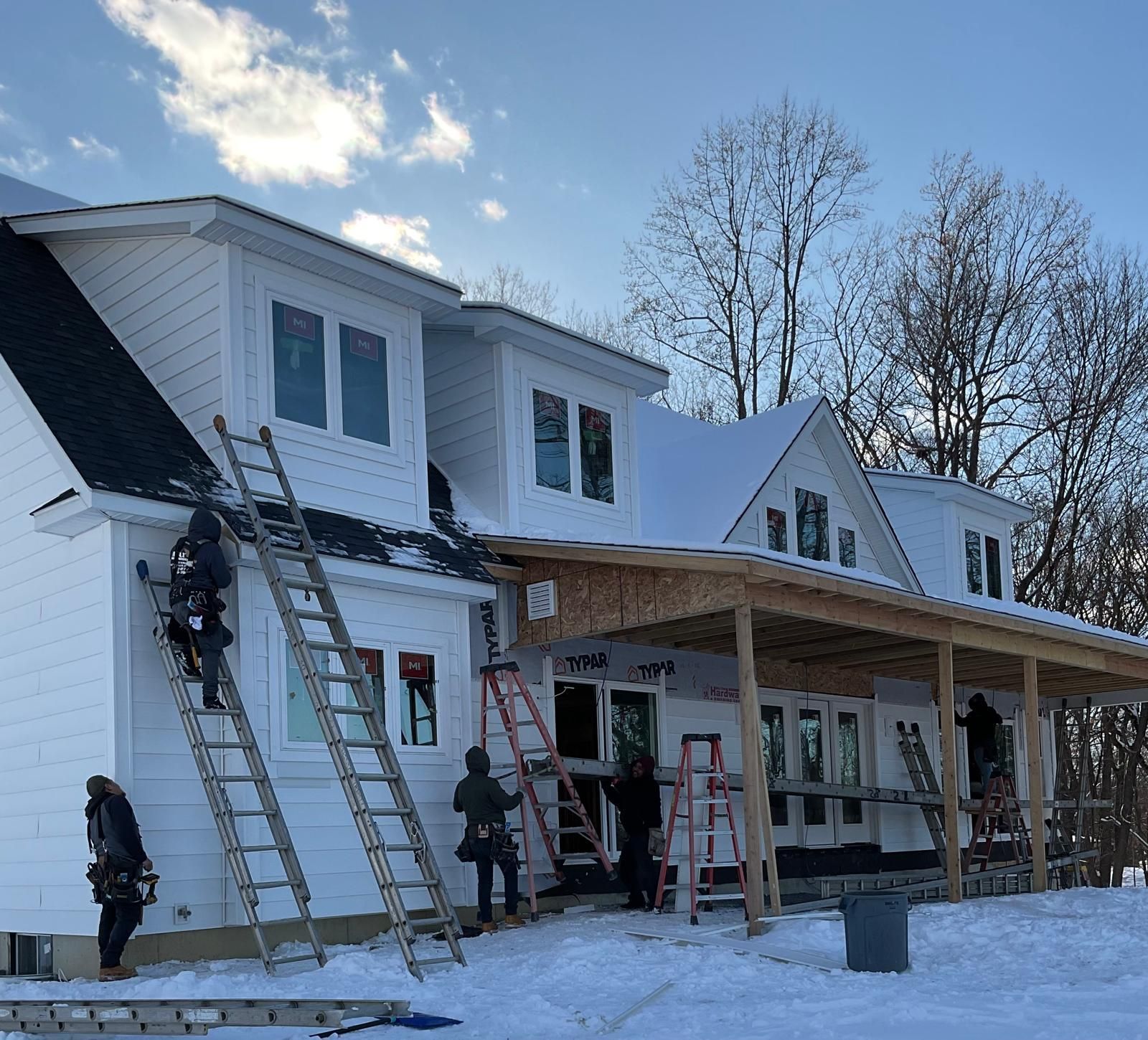 Construction workers on ladders and scaffolding install siding on a white house during winter with snow on the ground.