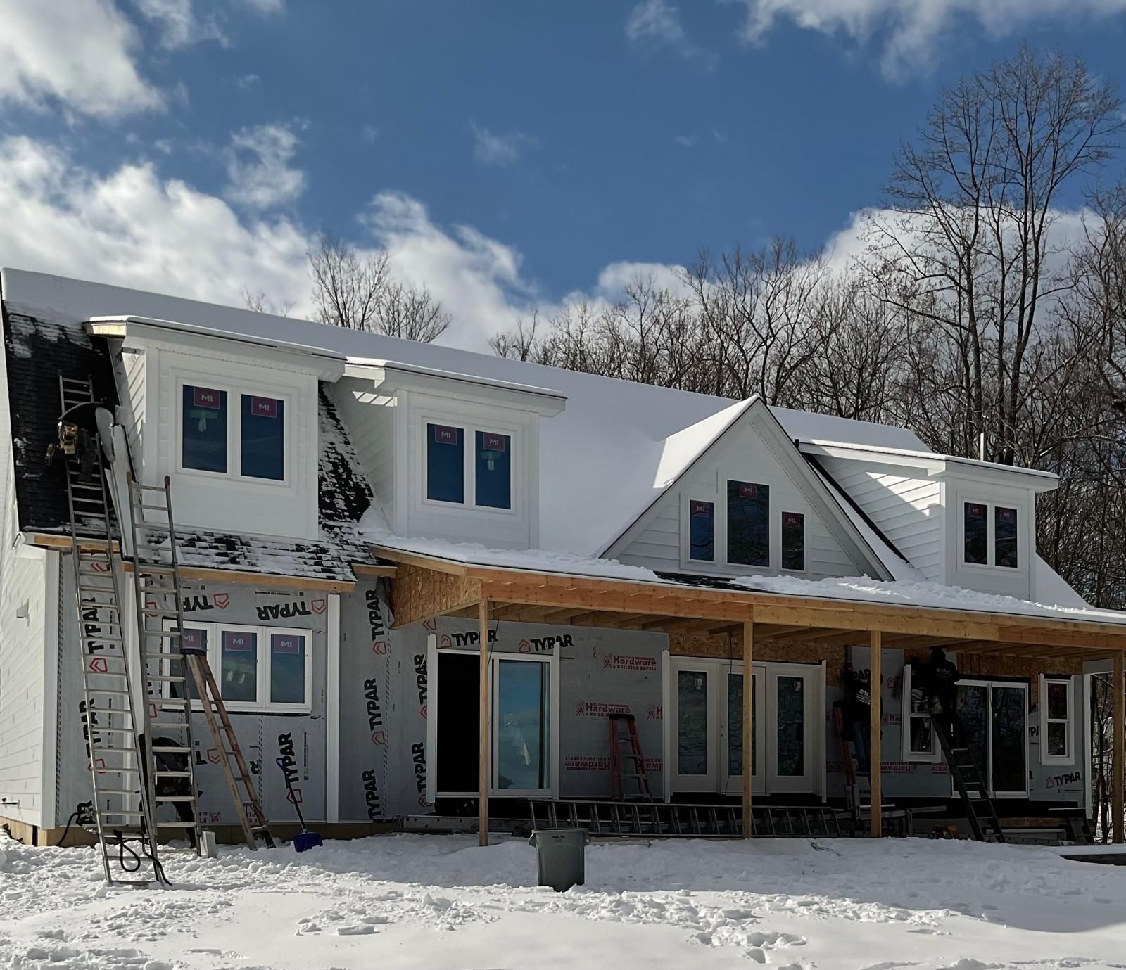A house under construction with a snowy roof, exposed framing on the porch, and scaffolding visible on the left side.