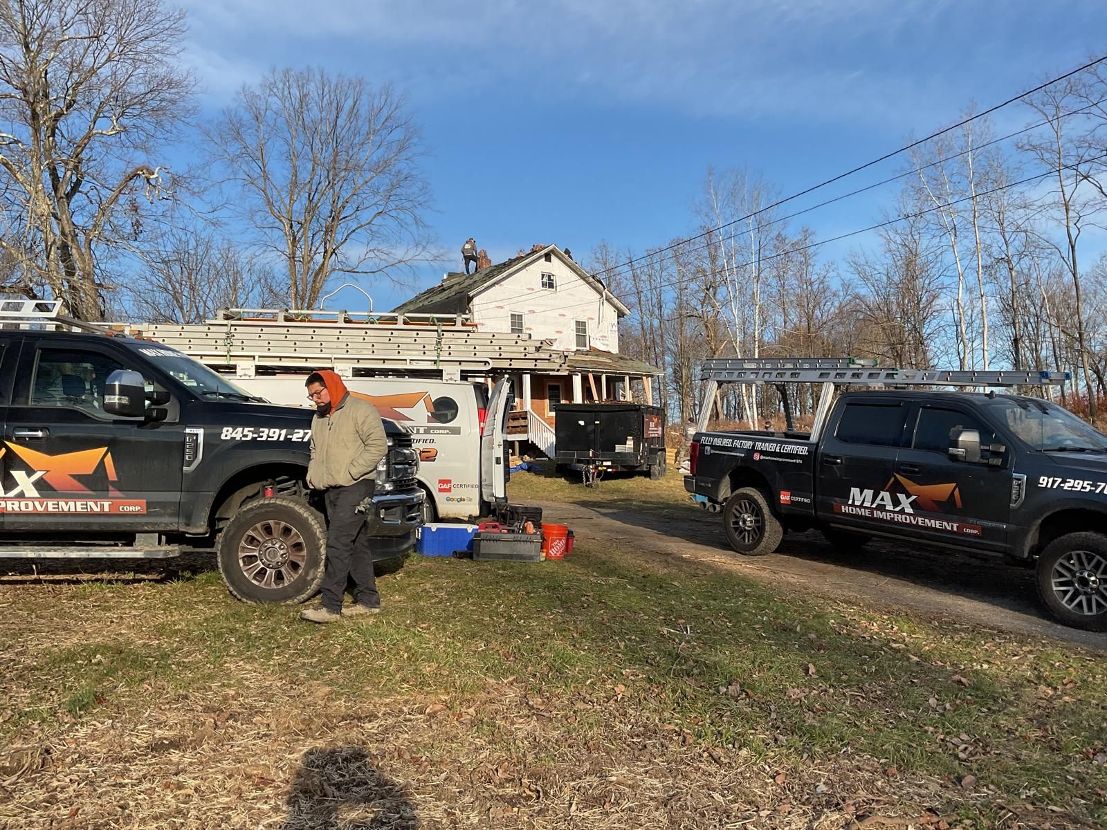 Two work trucks parked in a grassy field in front of an old two-story house under a blue sky.