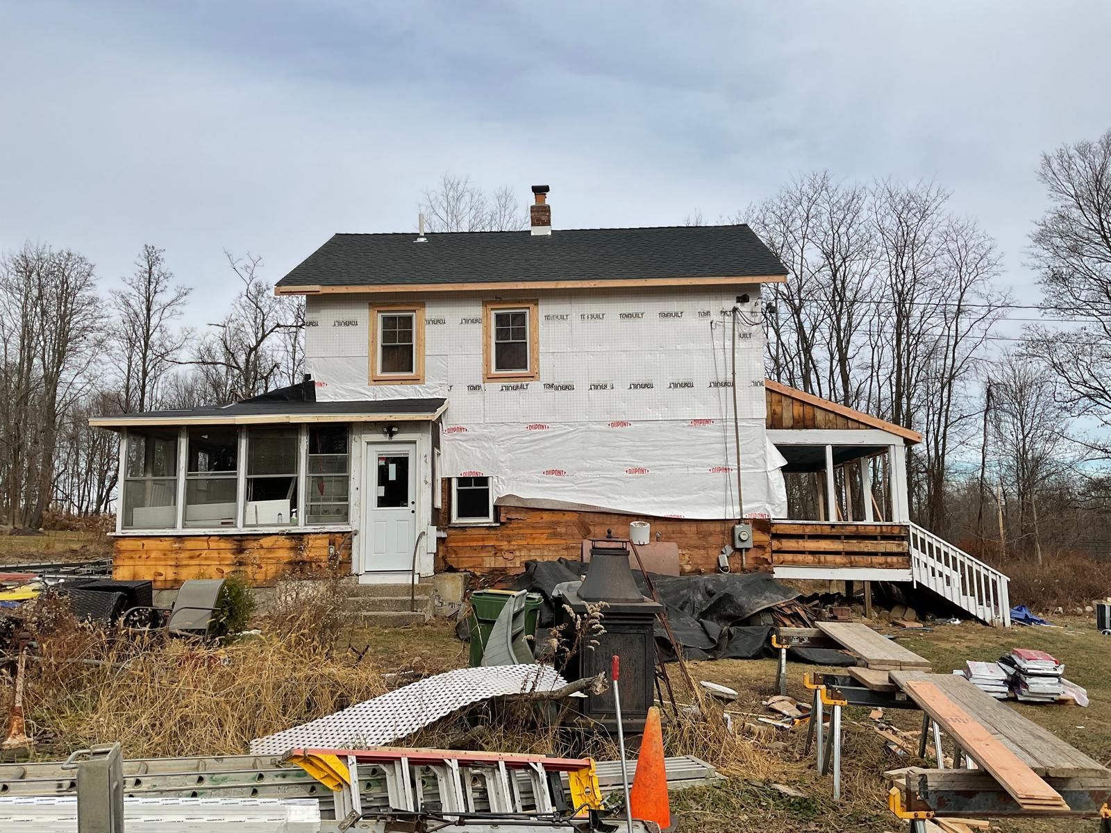 A two-story house under renovation with white house wrap, exposed wood framing, and construction debris in the yard.