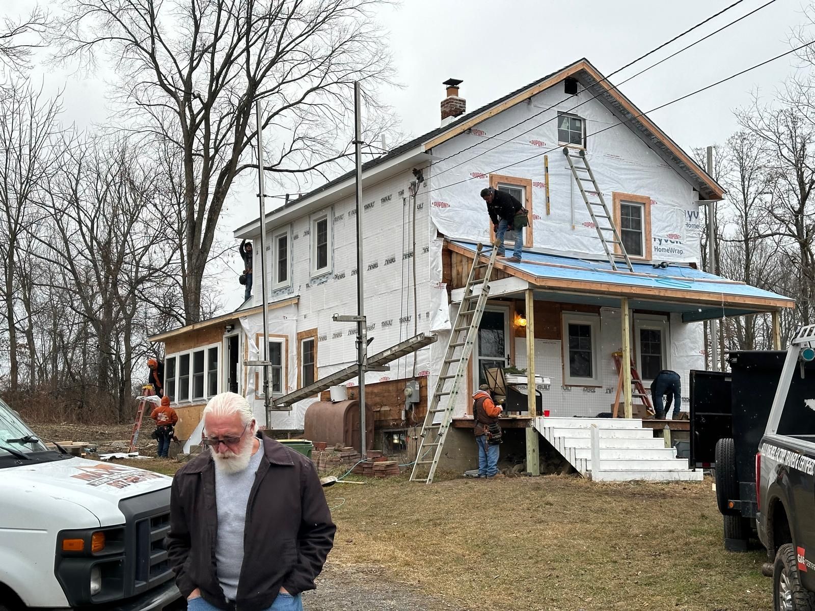 Workers renovate the exterior and roof of a two-story house while a person stands in the foreground.