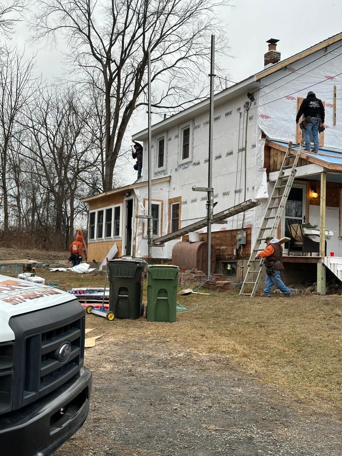Construction workers renovate the exterior of a two-story home covered in white house wrap, with a ladder and scaffolding.