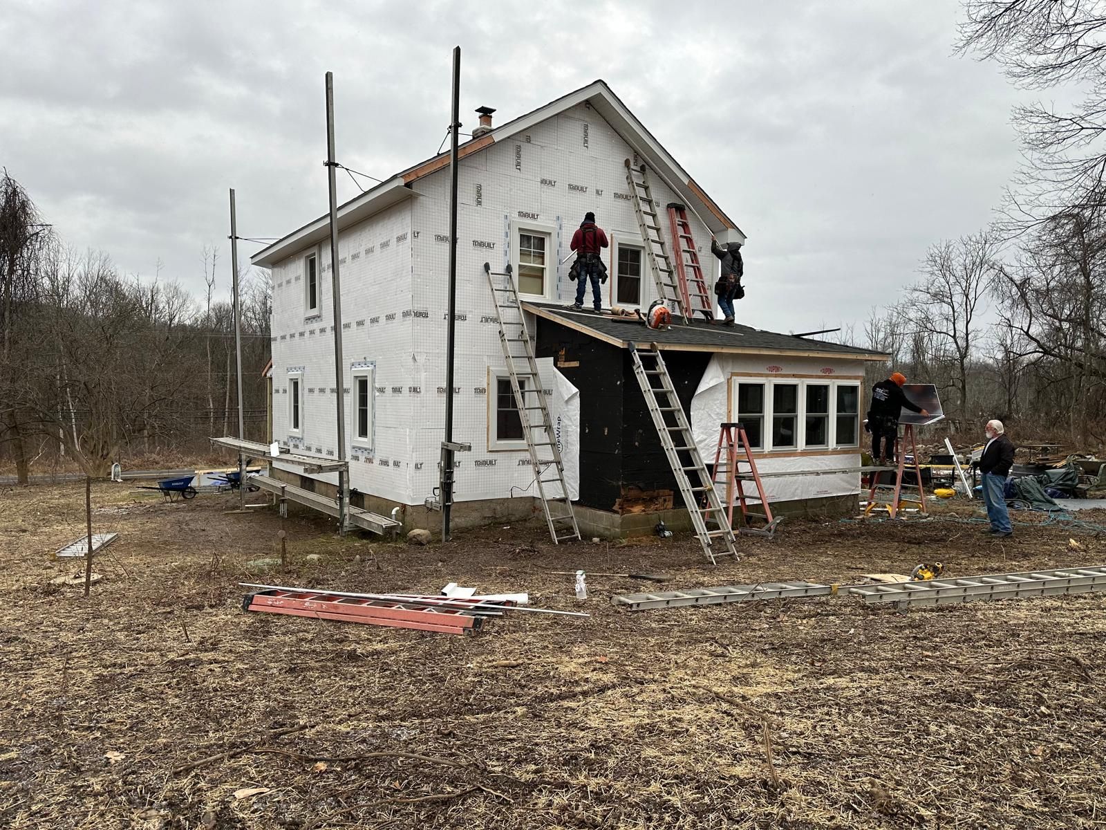 Workers on ladders renovate the exterior of a white, two-story house under a cloudy sky in a rural, wooded setting.